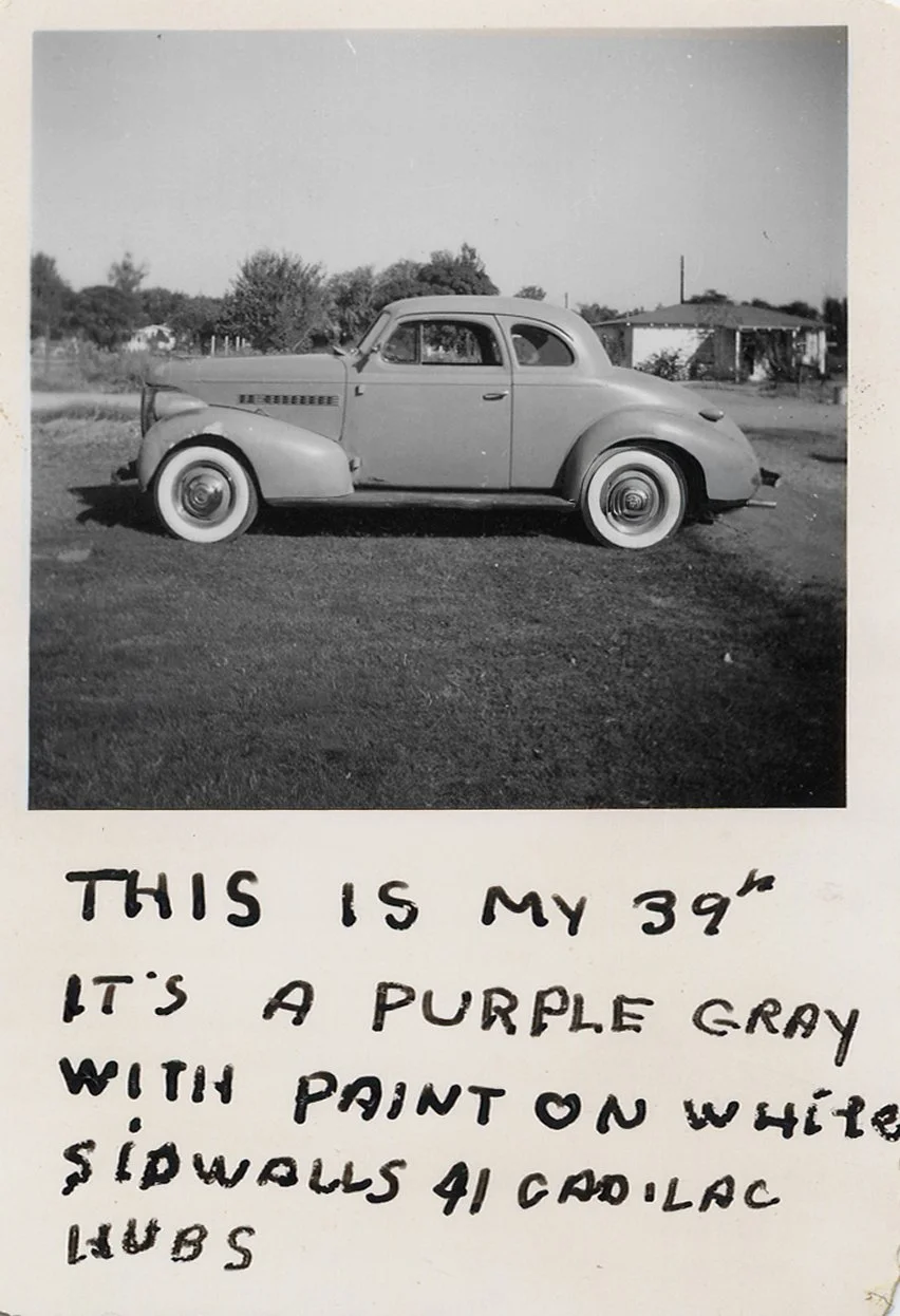 Black and white photo of a vintage car parked on a dirt lot with trees and buildings in the background; handwritten text underneath reads: "This is my 39 Oldsmobile. Its a purple gray with paint on white sidwalls 41 Cadillac hub caps."