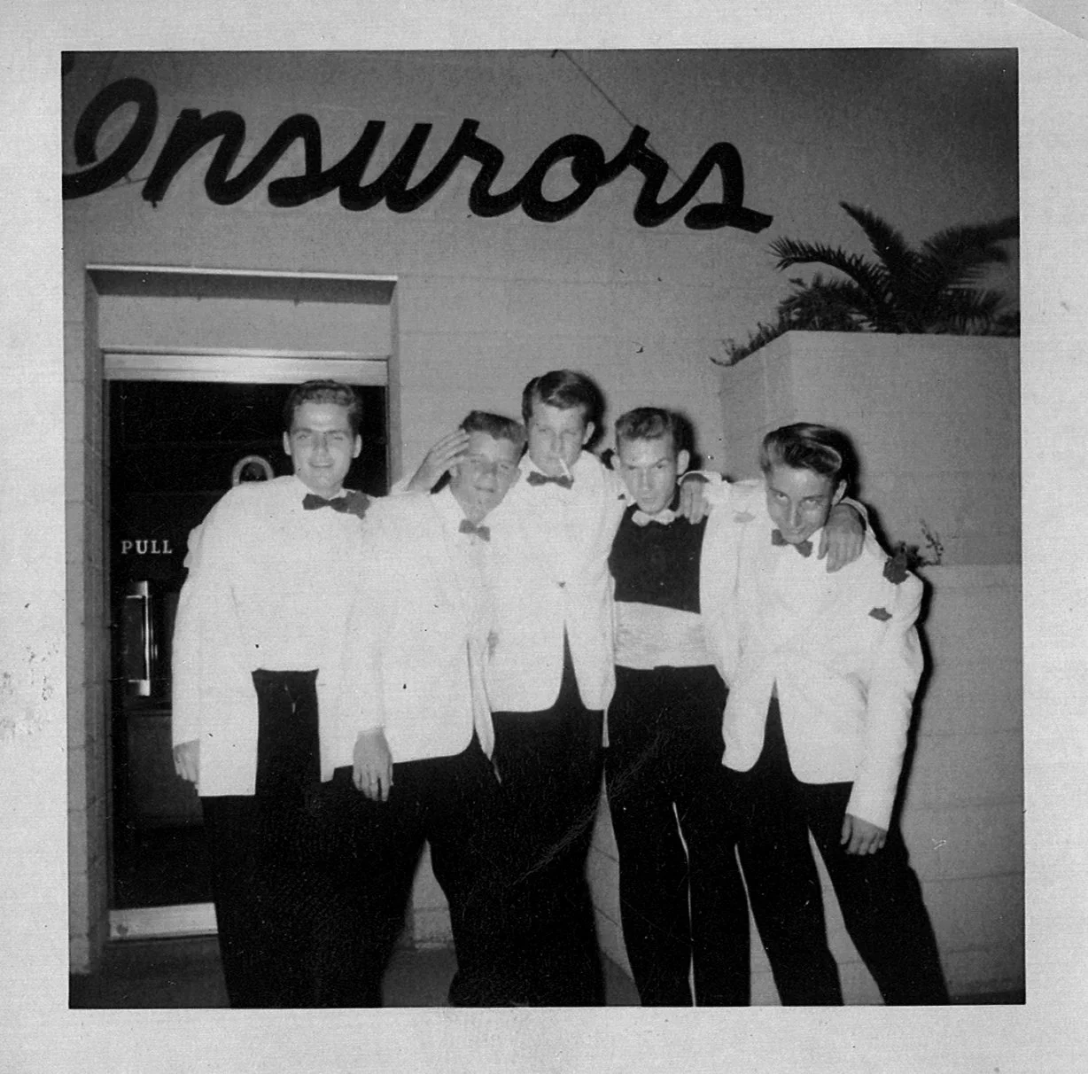 Black and white photo of five young men dressed in tuxedos with bow ties, standing close together with arms around each other, outside a building with palm plants and a sign that appears to spell 'Sonnose'. My Dad.