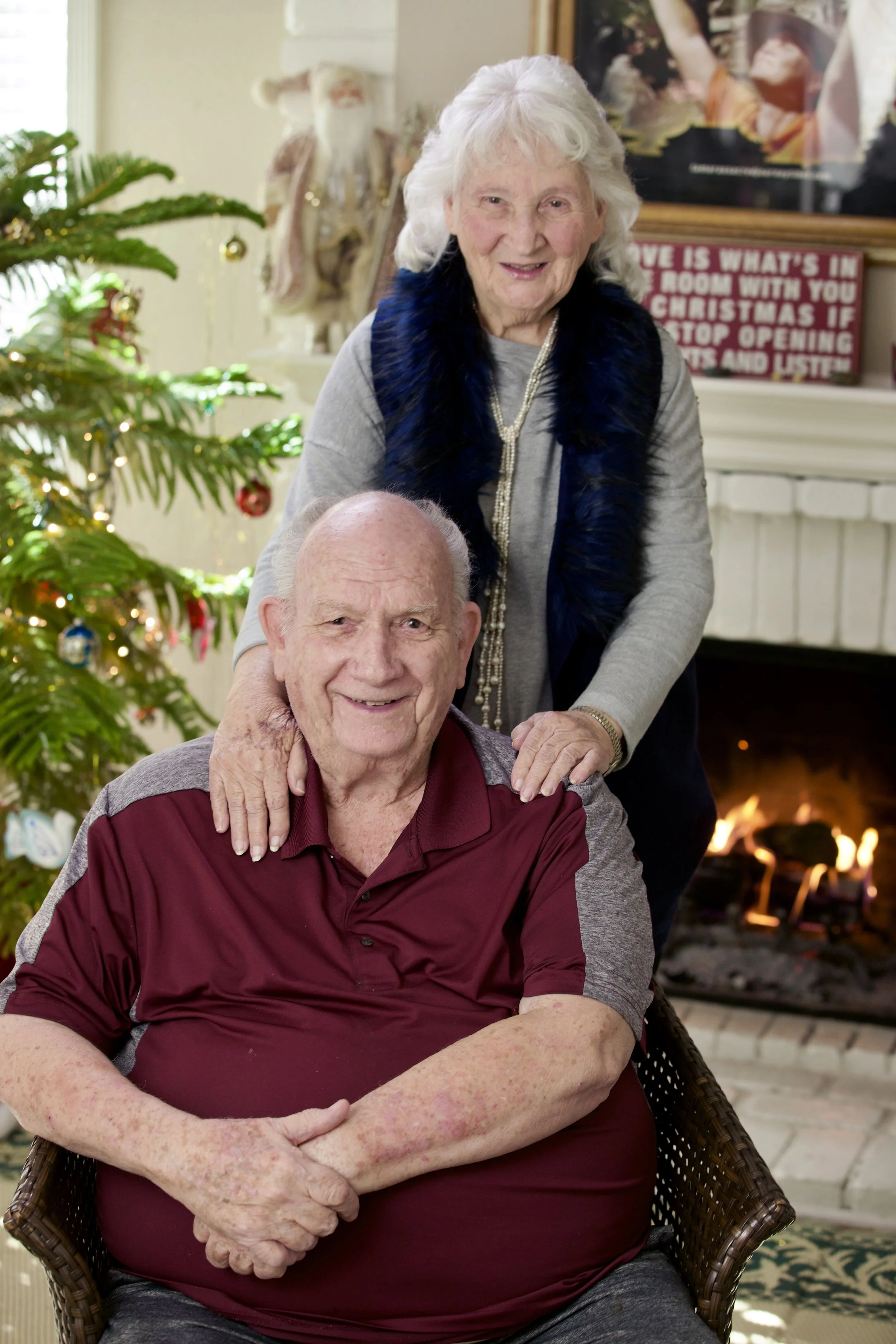 An elderly man and woman smiling indoors during Christmas. The man is seated in a wicker chair wearing a maroon polo shirt, and the woman is standing behind him with her hands on his shoulders, wearing a gray top and a dark fur necklace. There is a decorated Christmas tree with lights and ornaments to the left. In the background, a fireplace with a sign that reads 'Love is what's in the room with you at Christmas if you stop opening presents and listen,' an elf decoration, and a framed picture are visible. A fire is burning in the fireplace.
