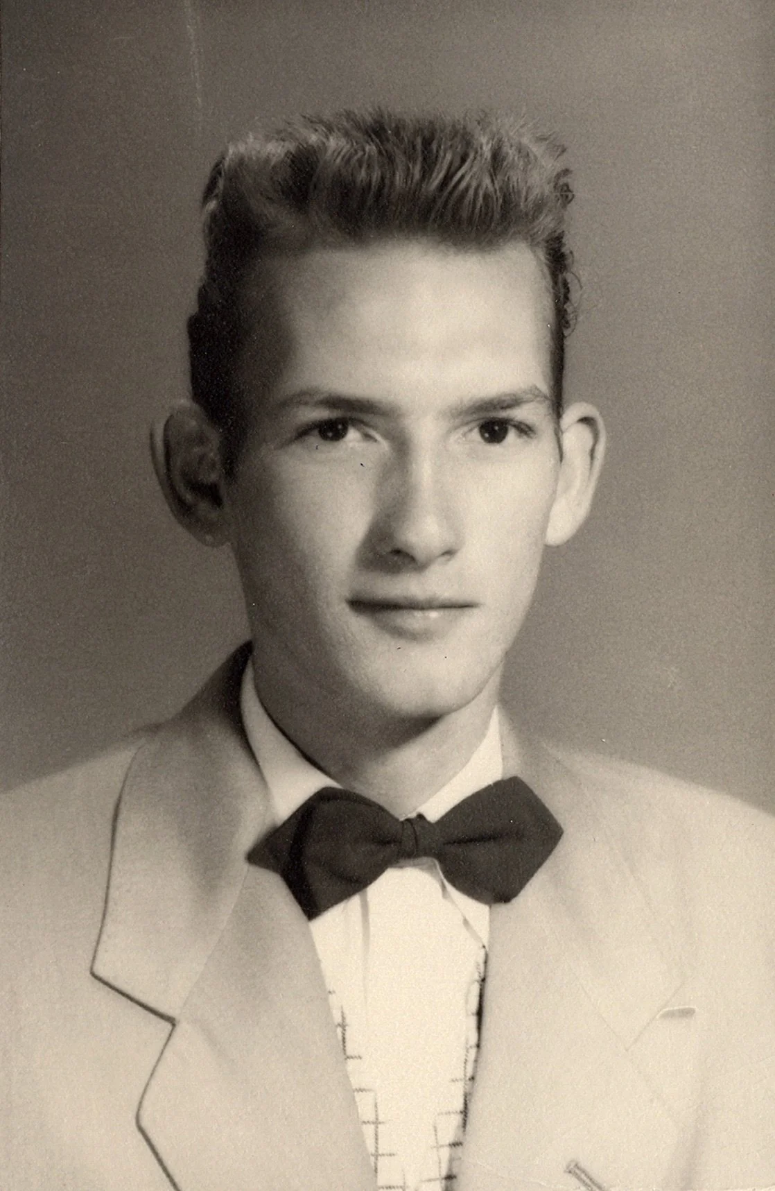Black and white portrait of a young man dressed in a tuxedo with a bow tie, with a light-colored suit jacket and shirt, smiling slightly at the camera. My Dad at 18.