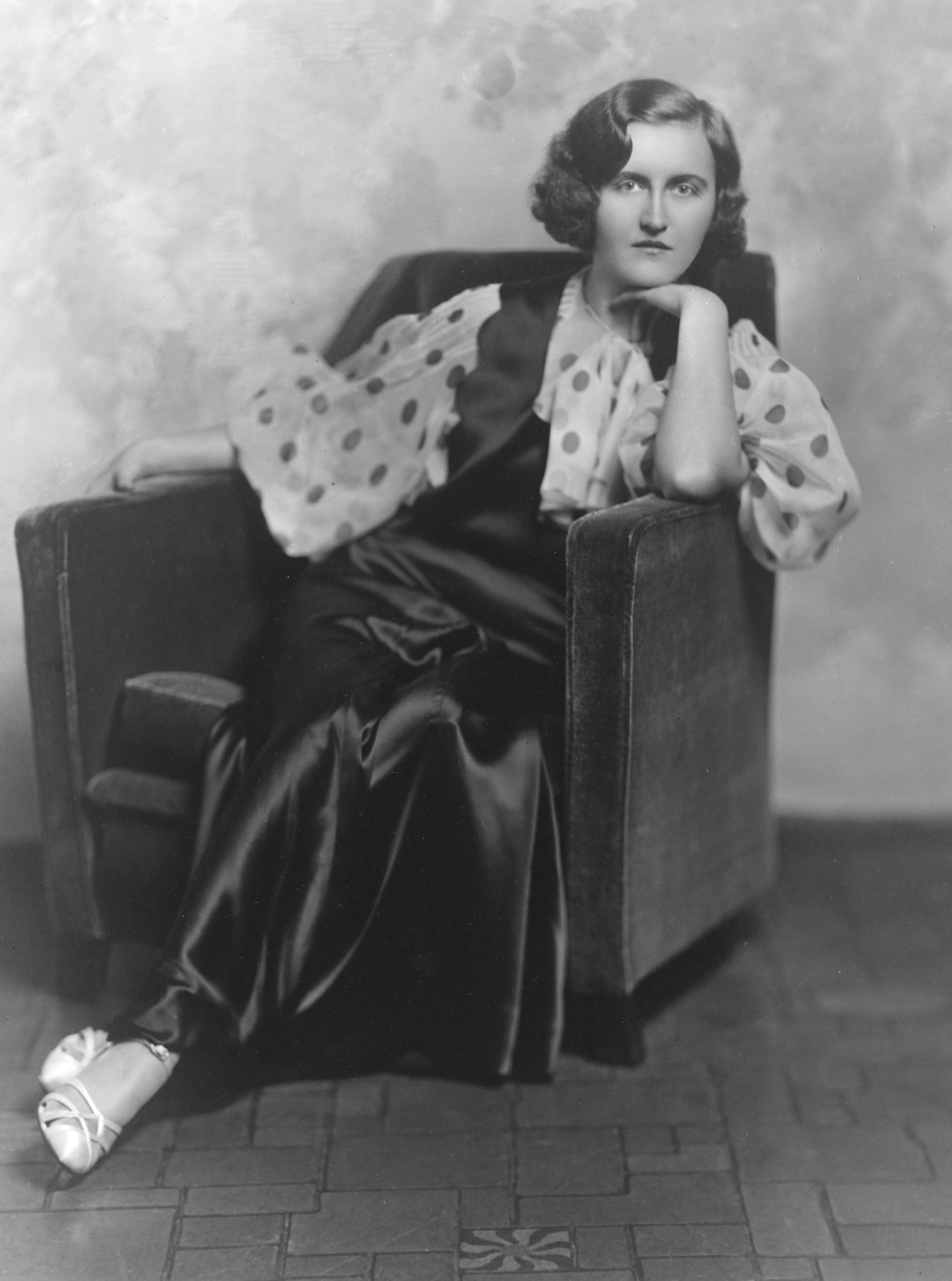Black and white photo of a woman sitting on a chair with her chin resting on her hand. She wears a polka dot blouse, a dark vest or cardigan, and shiny shoes. The setting appears to be indoors, with a tiled floor. Huguette Clark