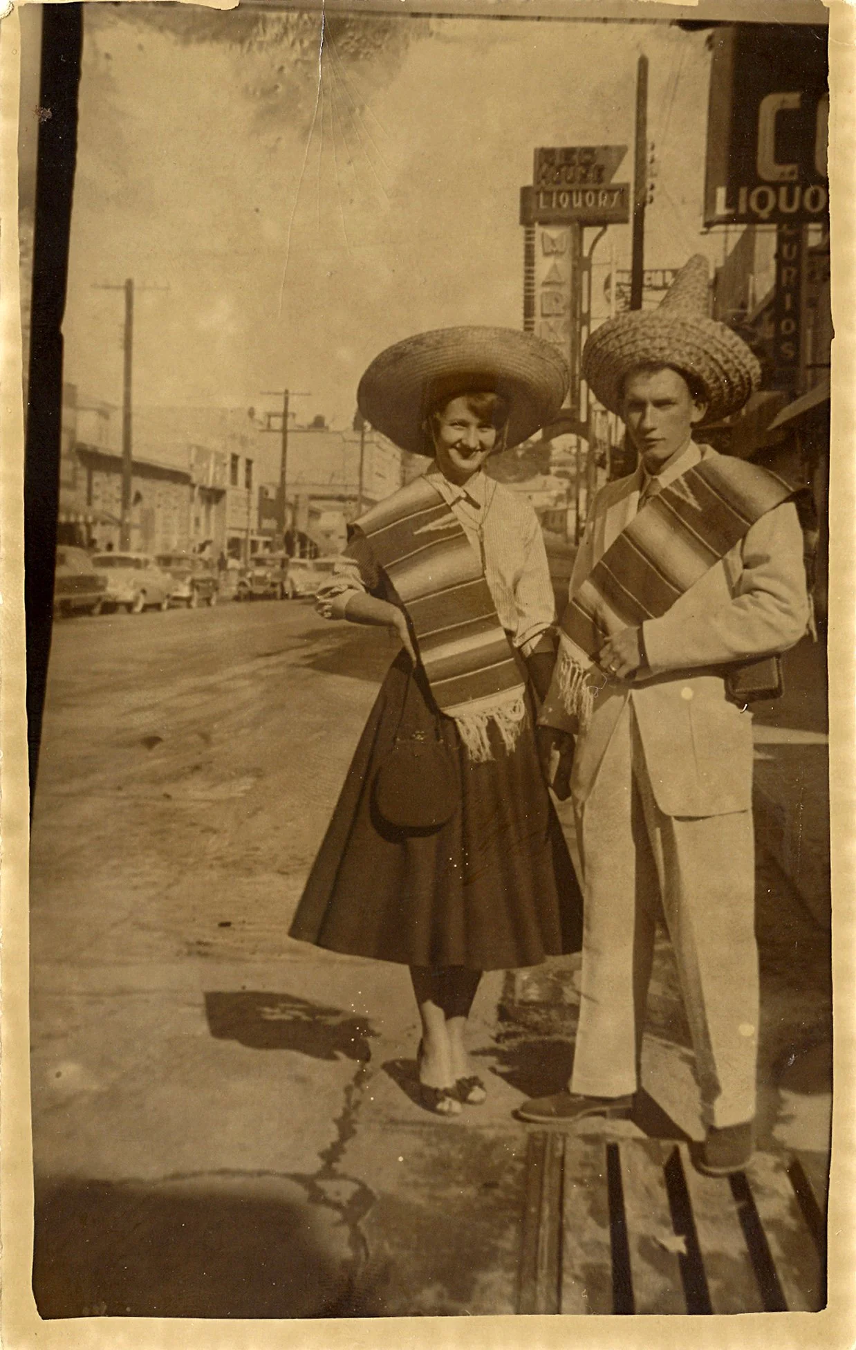 A vintage black-and-white photograph of a smiling woman and a serious man standing outside on a sidewalk. Both are wearing large, wide-brimmed sombrero hats and traditional Mexican garments. My Mom and Dad on their Honeymoon