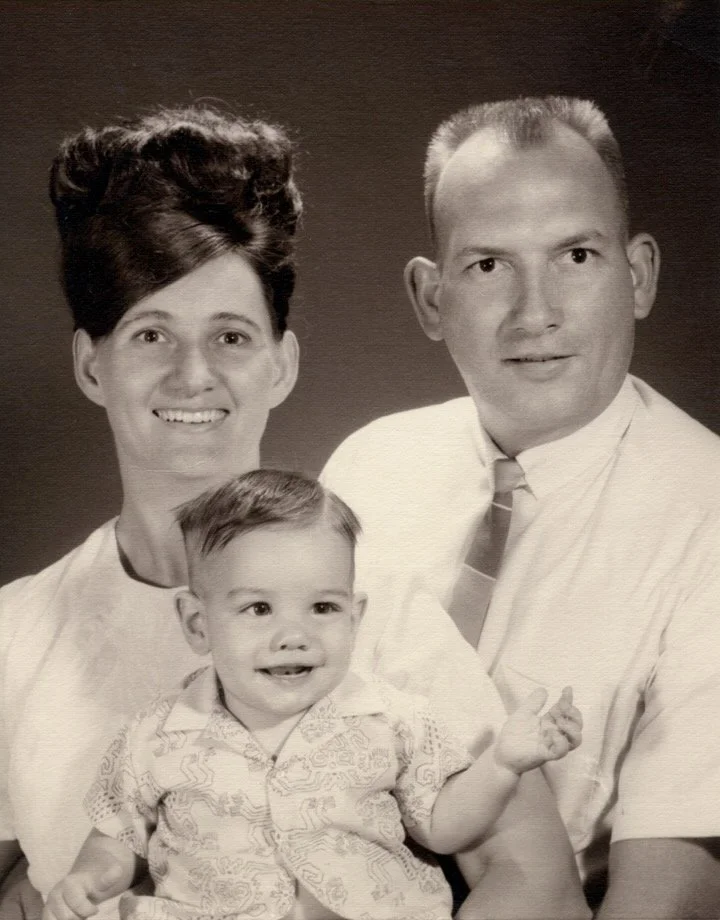 Black and white photograph of a smiling family portrait featuring a woman with short, dark hair, a man with short, light hair, and a young child with short, dark hair, all dressed in vintage-style clothing. Sumner Family ...Thats me in the Middle.