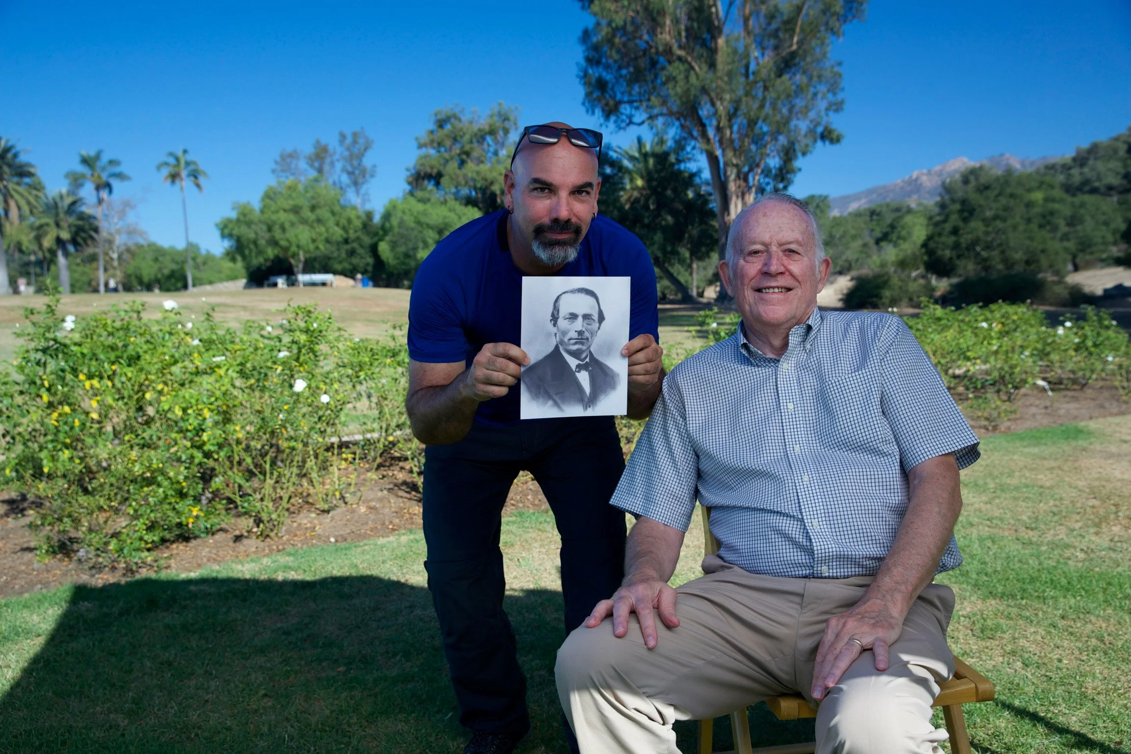 Two men outdoors, one seated and one standing. The man standing has a shaved head, goatee, sunglasses on his head, and is holding a black and white photograph of a man in a tuxedo. The seated man has short white hair, is smiling, and is wearing a checkered shirt. The background features trees, bushes, hills, and a clear blue sky.