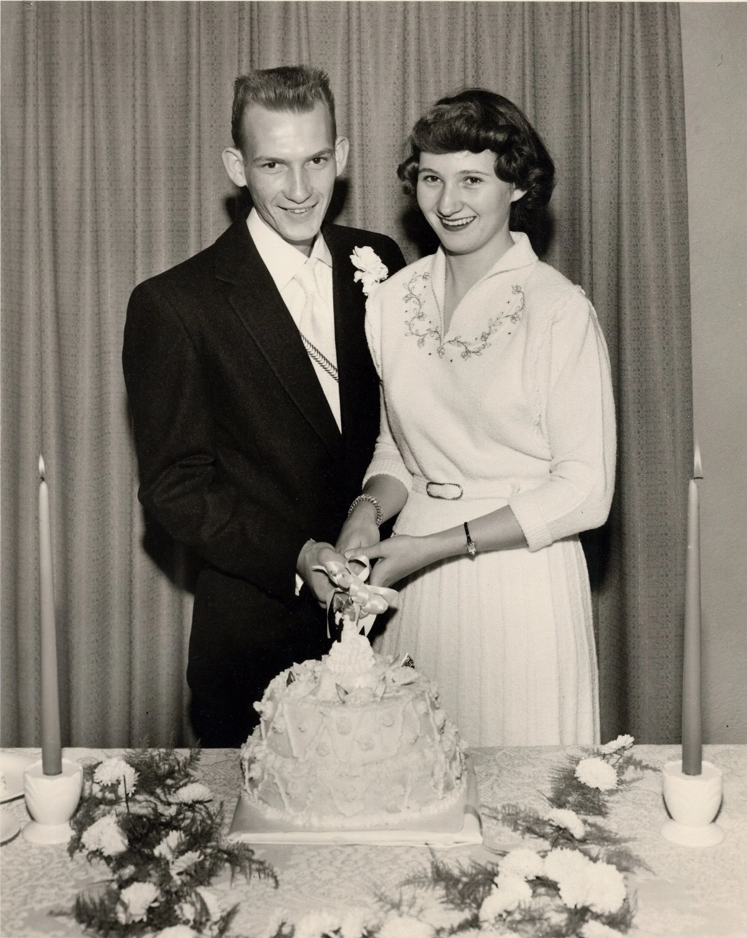 A black-and-white photo of a man and woman smiling and holding hands in front of a decorated table with a cake, flowers, and greenery, with two vertical poles and curtains in the background. My Mom and Dad's Wedding