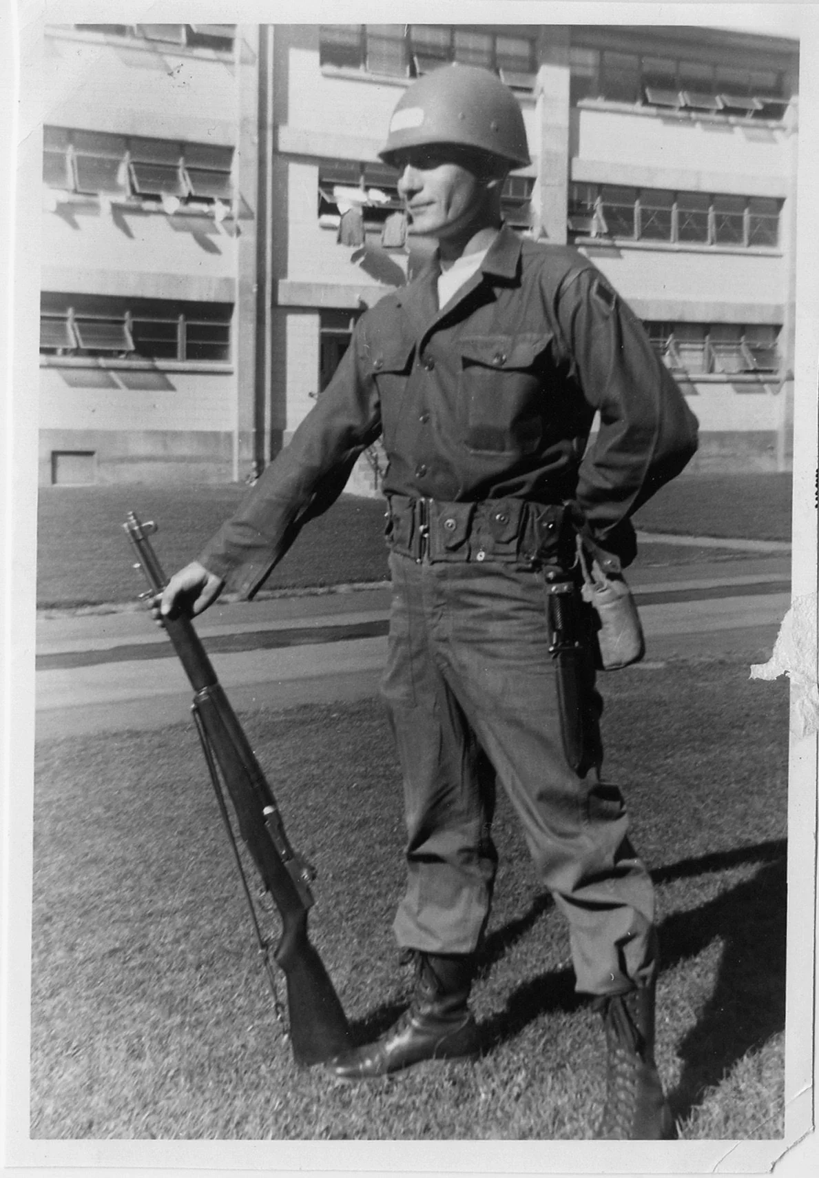 Black and white photograph of a soldier standing outdoors on a grassy area, wearing a helmet and military uniform, holding a rifle in one hand, with a modern multi-story building in the background.