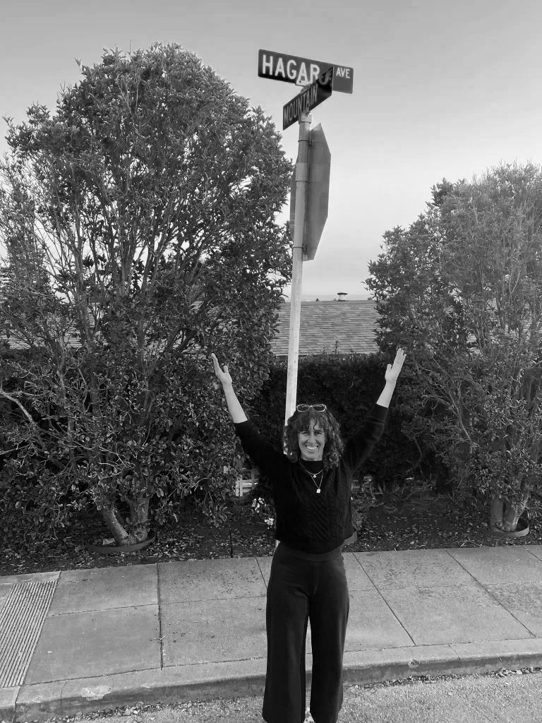 Writer Hagar Scher stands in front of a street sign with her name on it, Hagar Ave with arms raised in joyful pose.