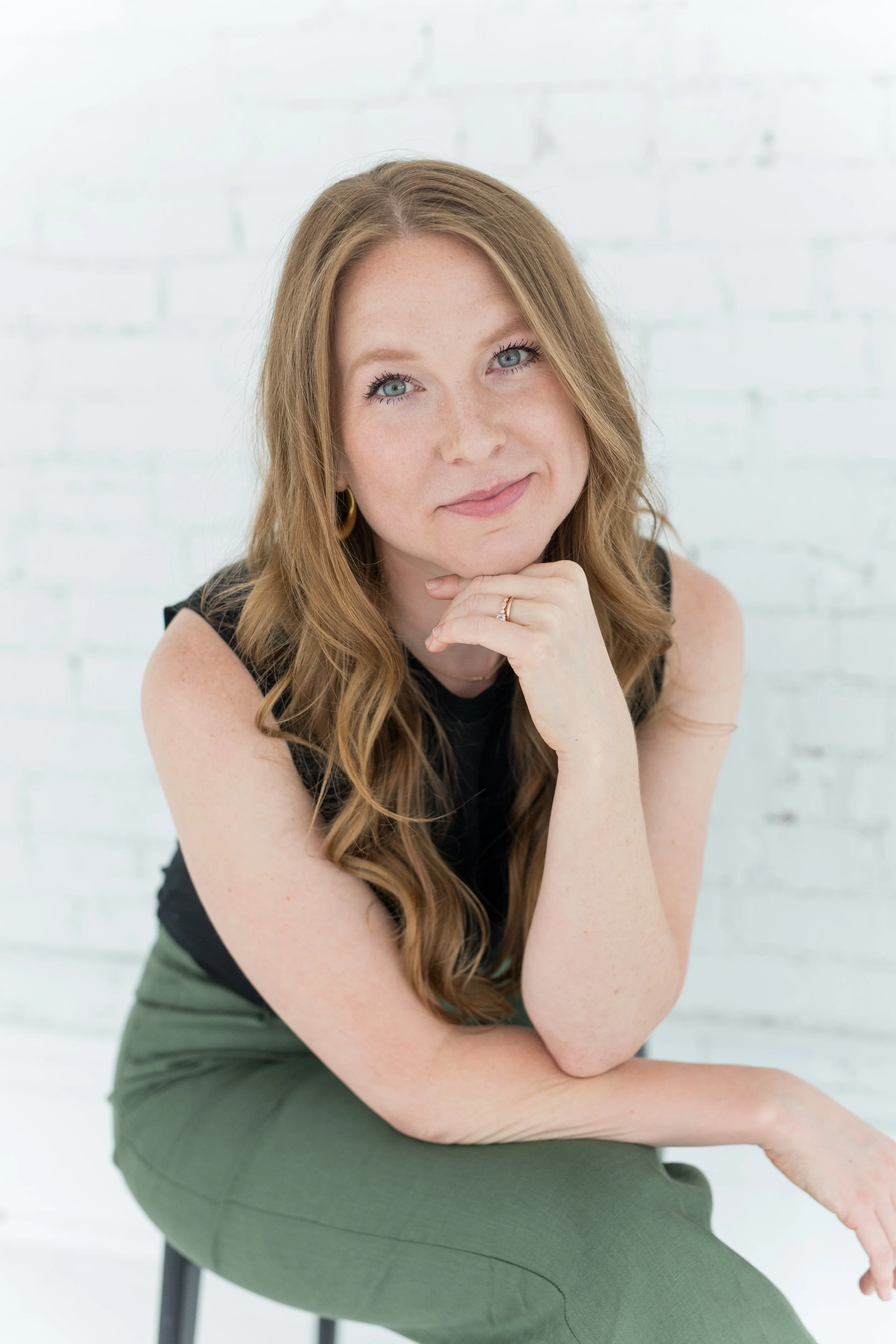 Portrait of a smiling woman with long red hair, blue eyes, resting chin on hand, wearing a black top and green pants, seated against a white brick wall.