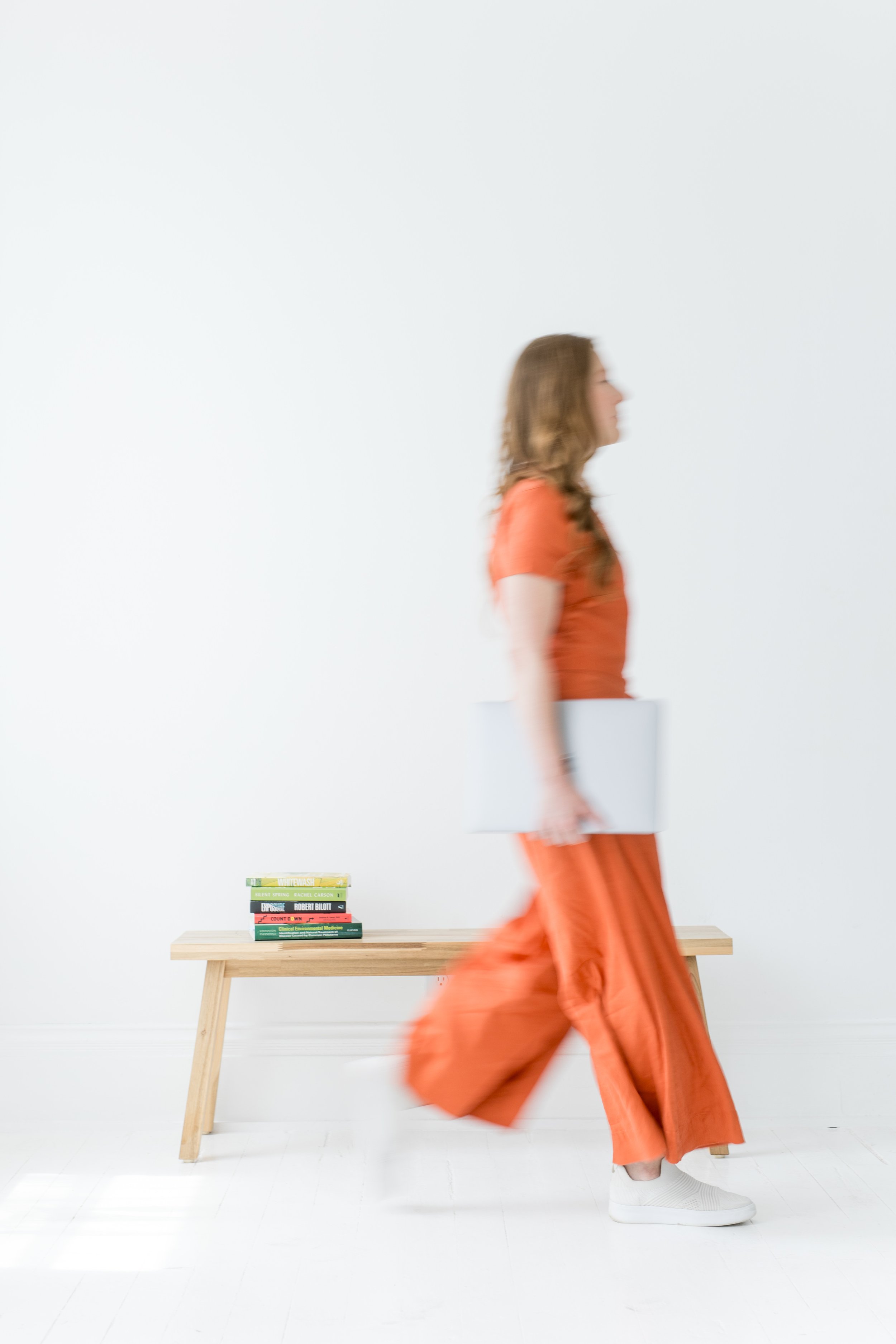 Person walking in orange outfit with white shoes in a minimalist room with a wooden table, a stack of colorful books, and a white wall.