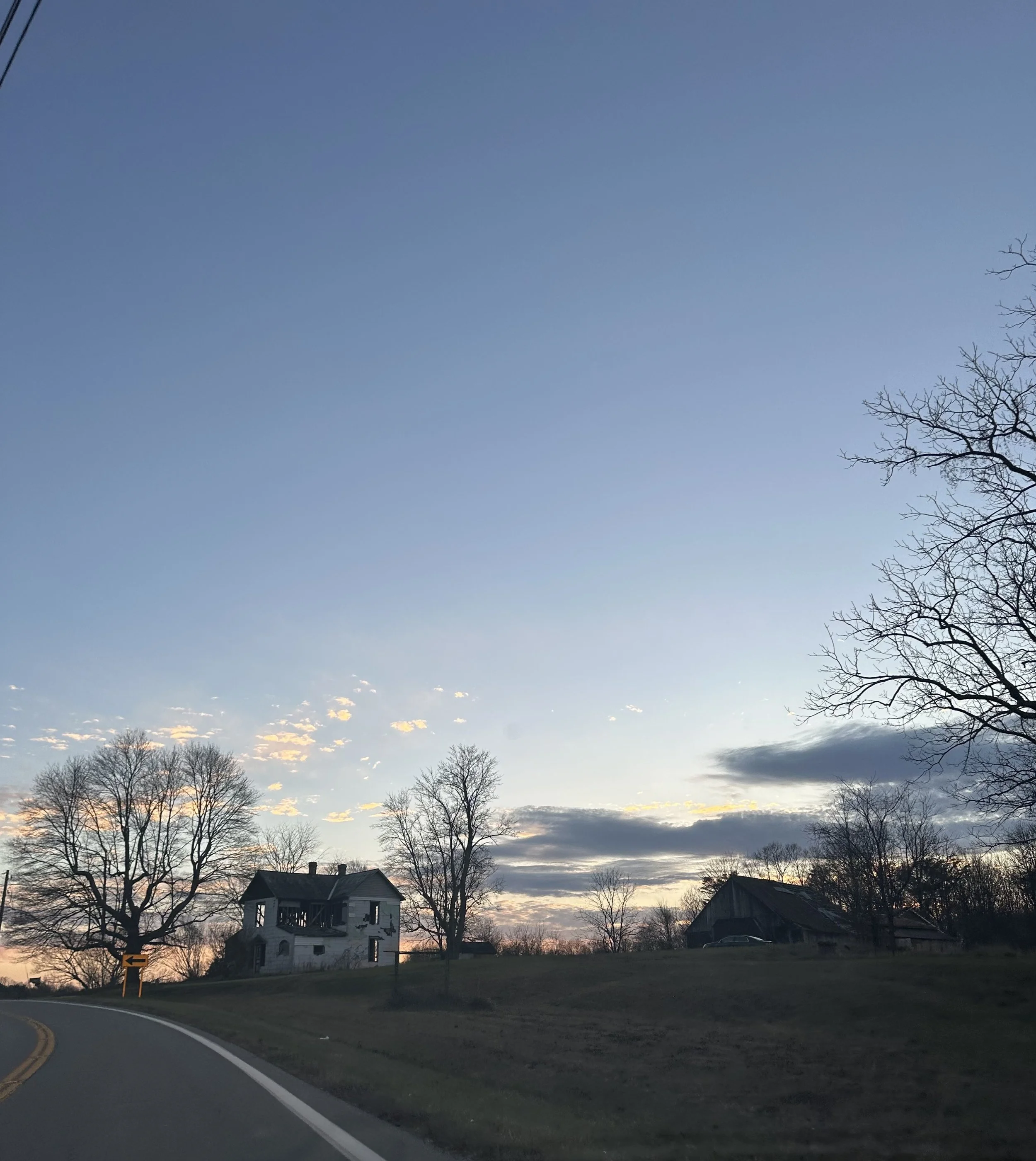 A winding road in a rural area at sunset or sunrise with leafless trees and a few houses in the background, some clouds in the sky, and a small road sign on the left side of the road.