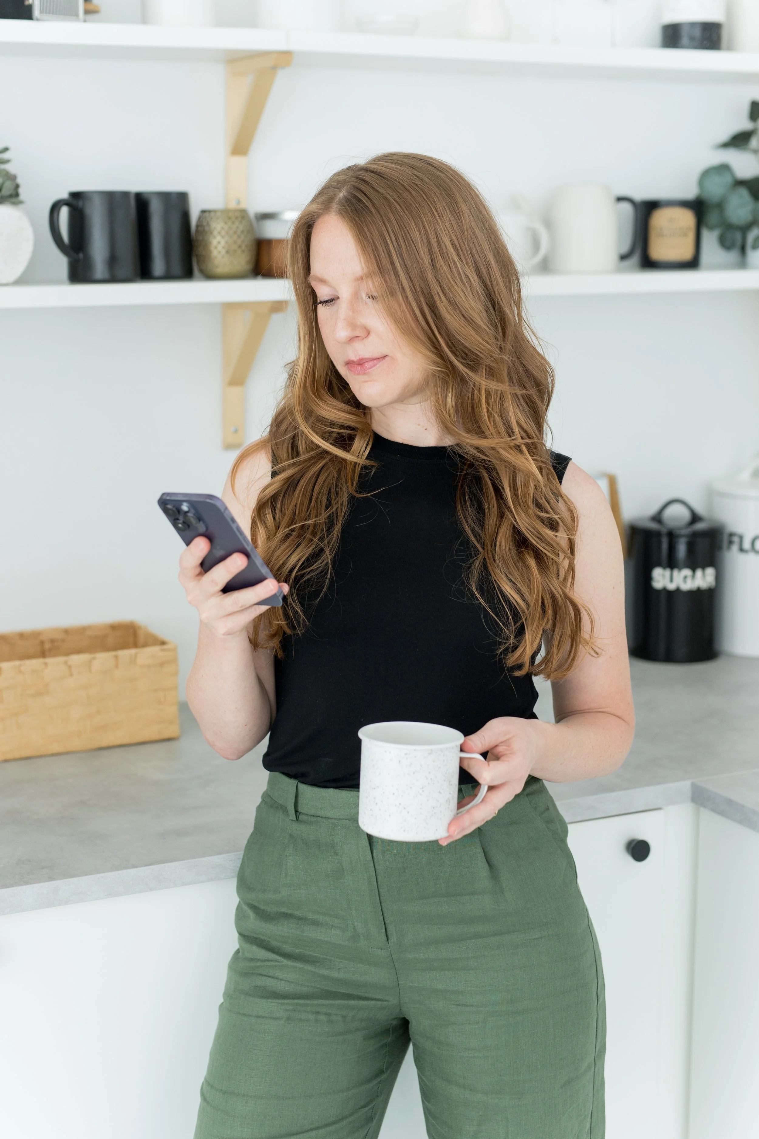 A woman with long reddish hair, wearing a black sleeveless shirt and green pants, holding a mug in one hand and looking at her phone in the other, standing in a modern kitchen.