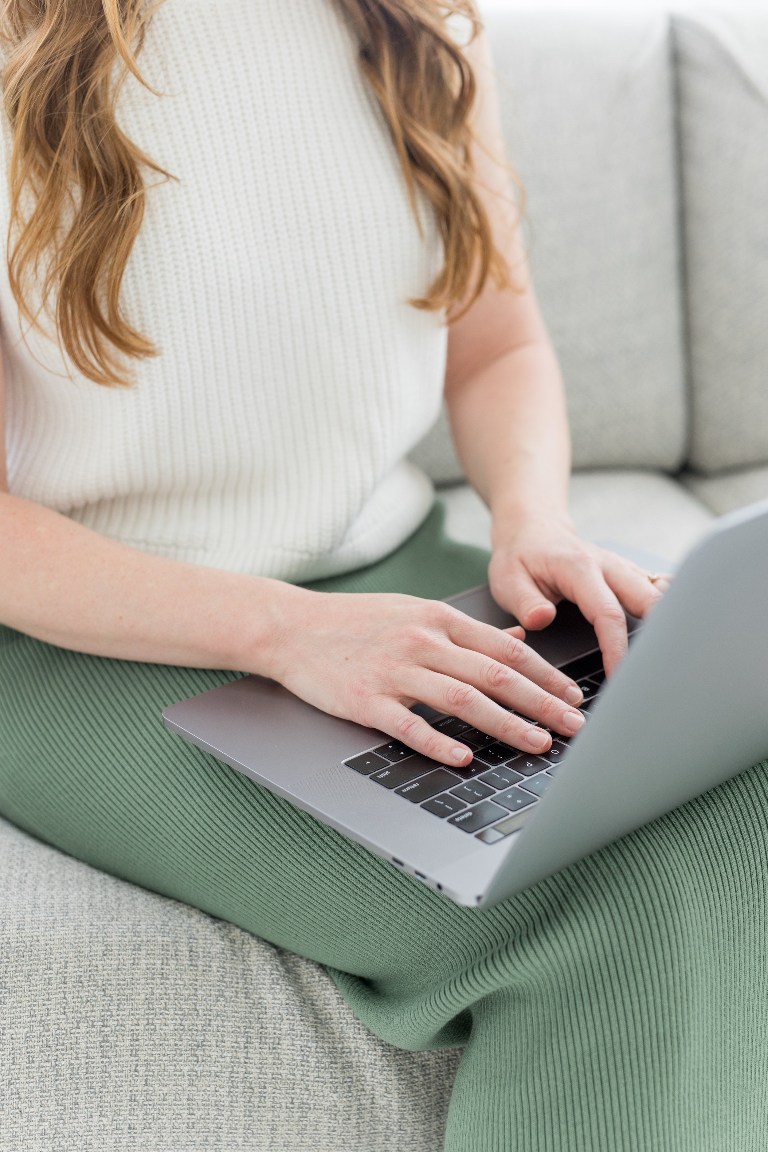 A woman with long, wavy, red hair is sitting on a light gray carpeted floor with a green yoga mat. She is wearing a white, short-sleeved, textured top and green pants. She is typing on a silver laptop.