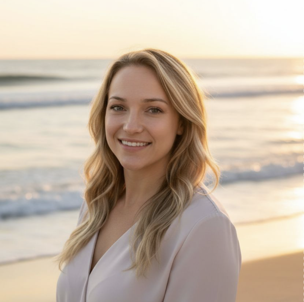A young woman with blonde hair smiling at the camera on a beach during sunset.