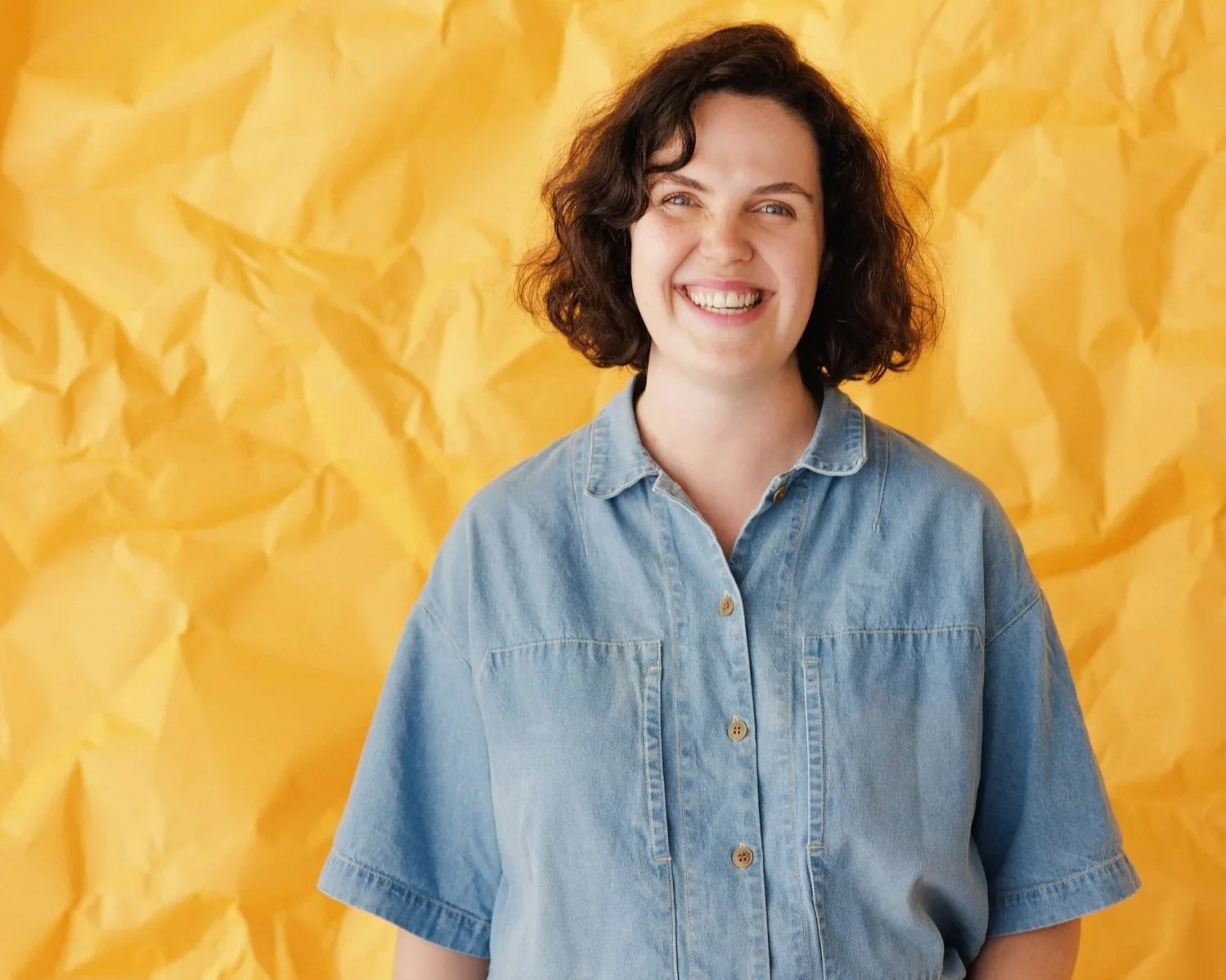 Brunette women smiles at camera in a blue denim top against a crumpled yellow backdrop