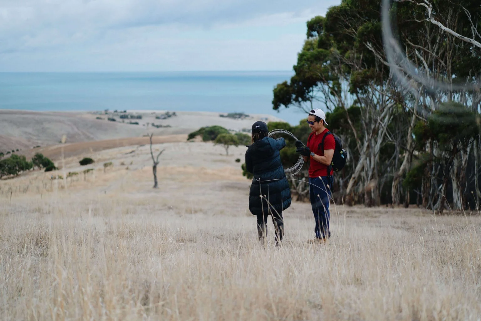 Two people preparing equipment in an open field near the coastline, with trees and a cloudy sky in the background.