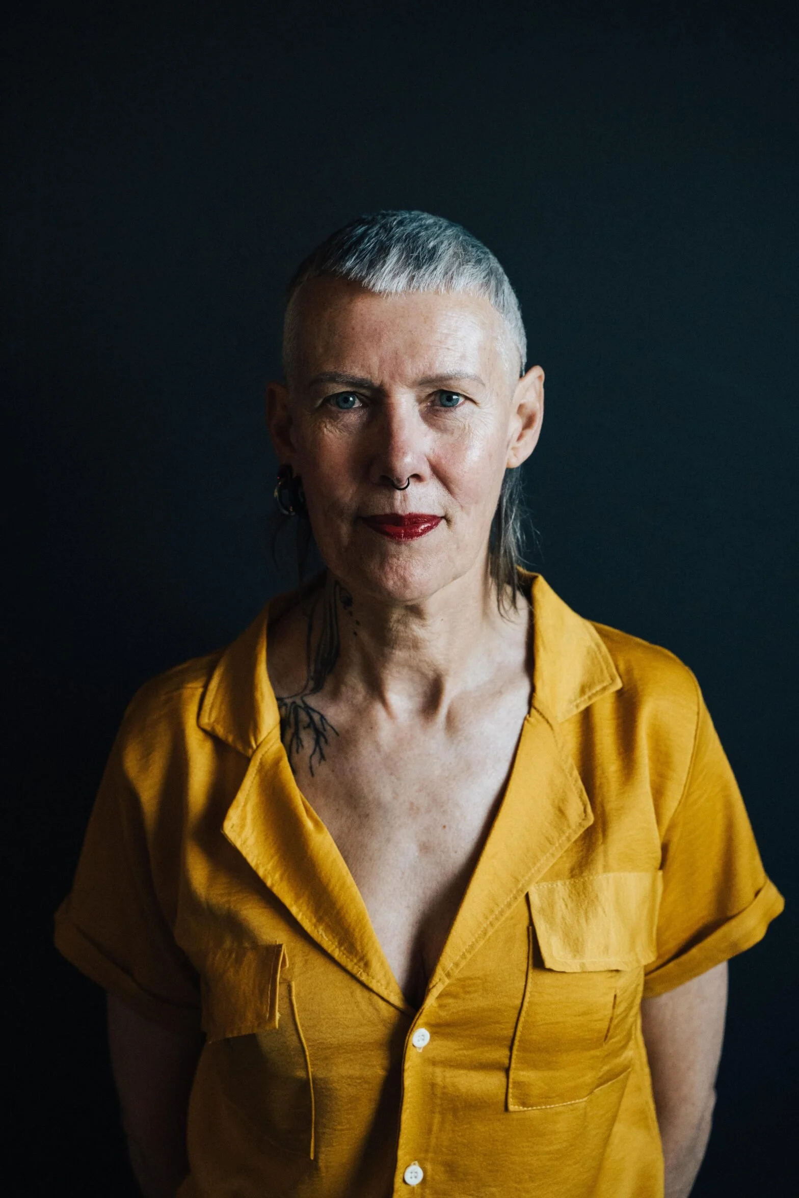 Portrait of a woman with short gray hair, blue eyes, and tattoos, wearing a yellow button-up shirt, against a dark background.
