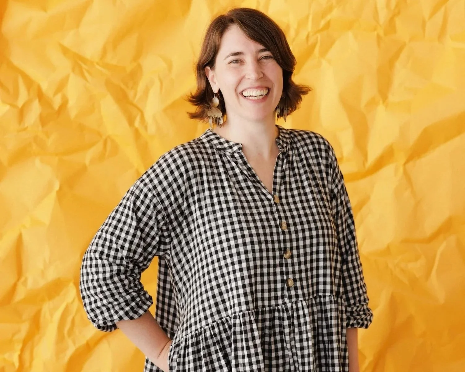 Women with brown smiles at the camera in a check black and white dress, against a crumpled yellow backdrop