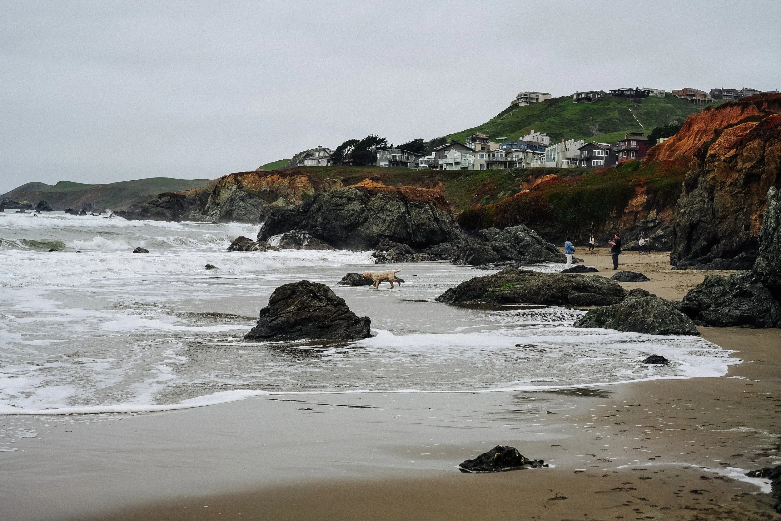 A coastal scene with waves crashing on a sandy beach, large rocks, and people walking along the shore. Houses are perched on a hillside in the background under a cloudy sky.