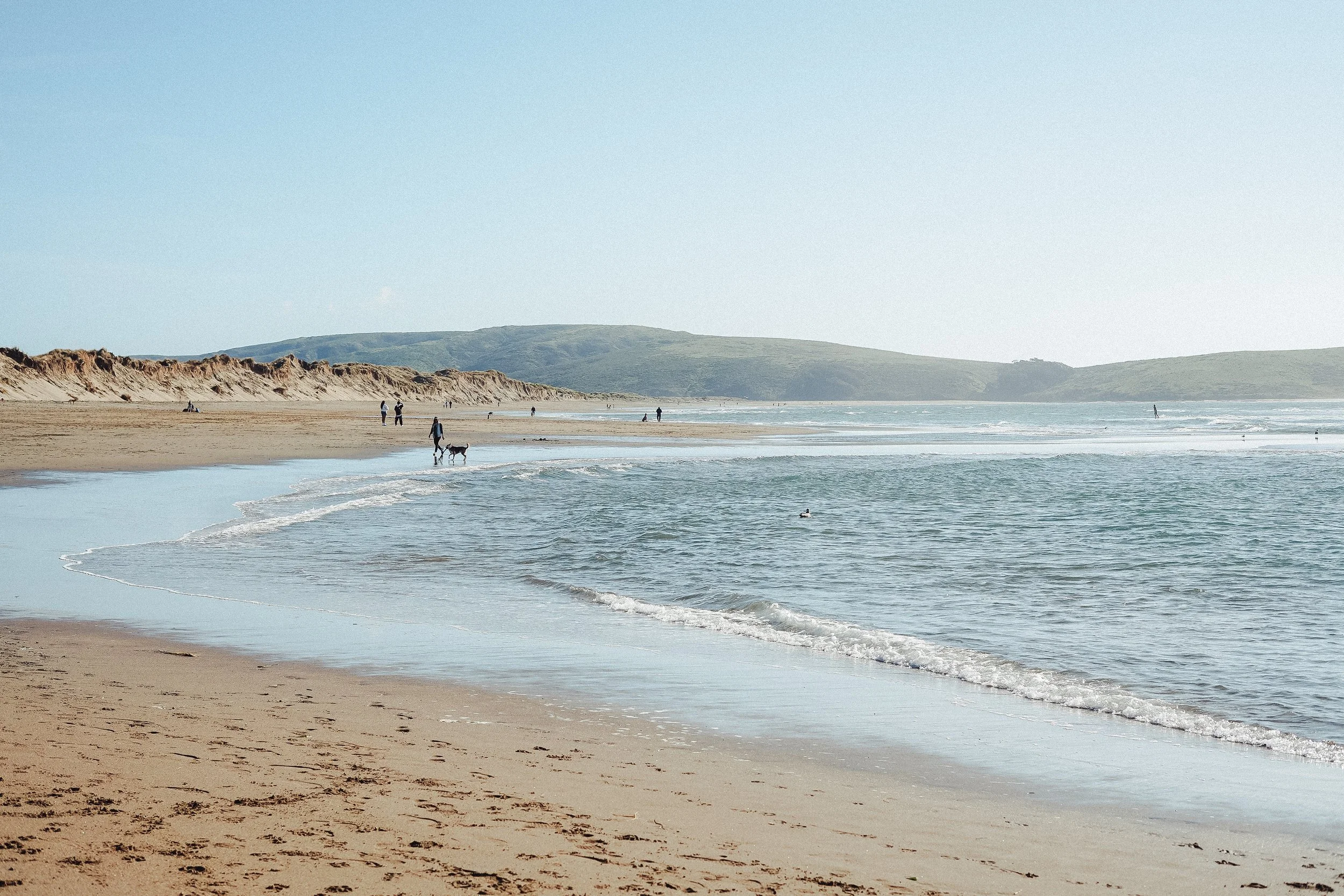 Beach with sandy shoreline, gentle waves, and a few people walking or relaxing, with hills in the background under a clear blue sky.