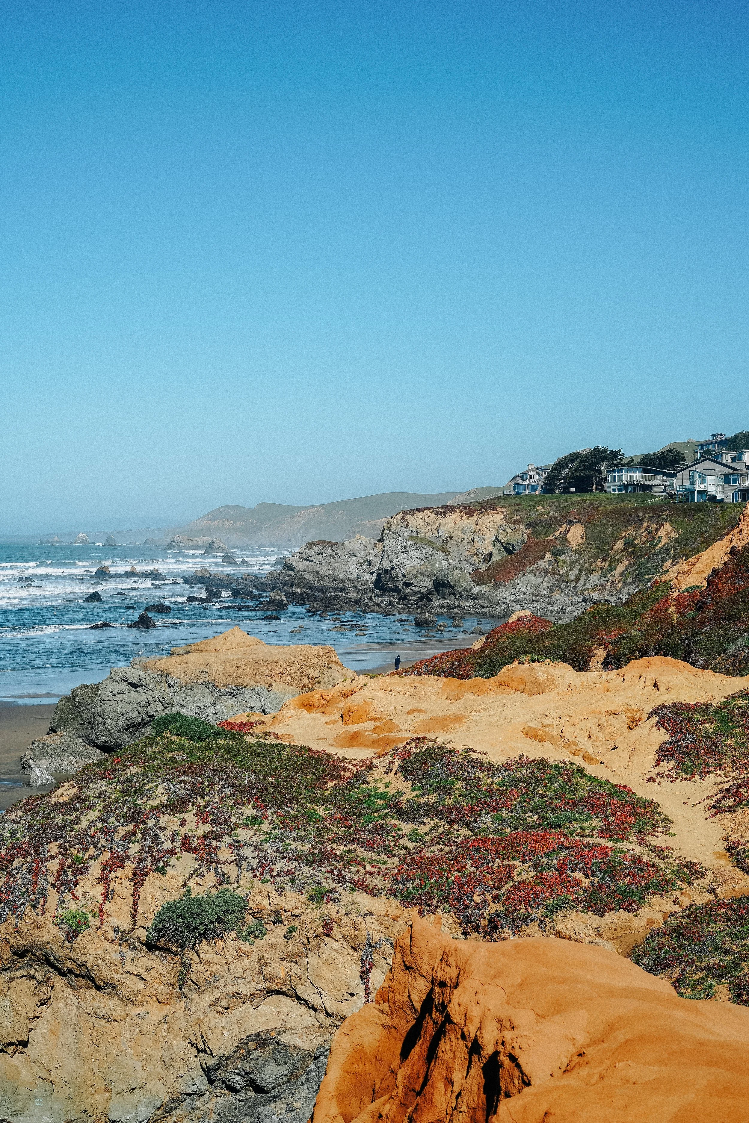 Coastal cliffs with houses on top, rocky shoreline, ocean waves, and a clear blue sky.