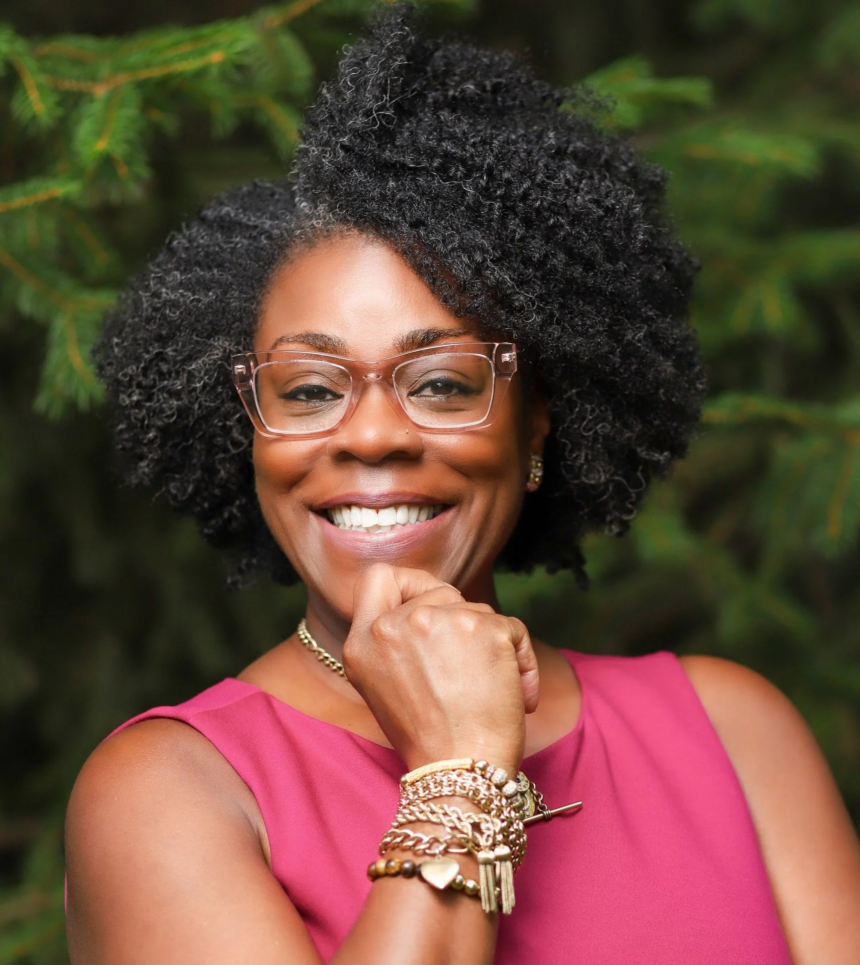 Close-up of an African American woman with curly black hair, wearing glasses, a pink top, and multiple gold bracelets, smiling with her hand near her chin, against a blurred green foliage background.