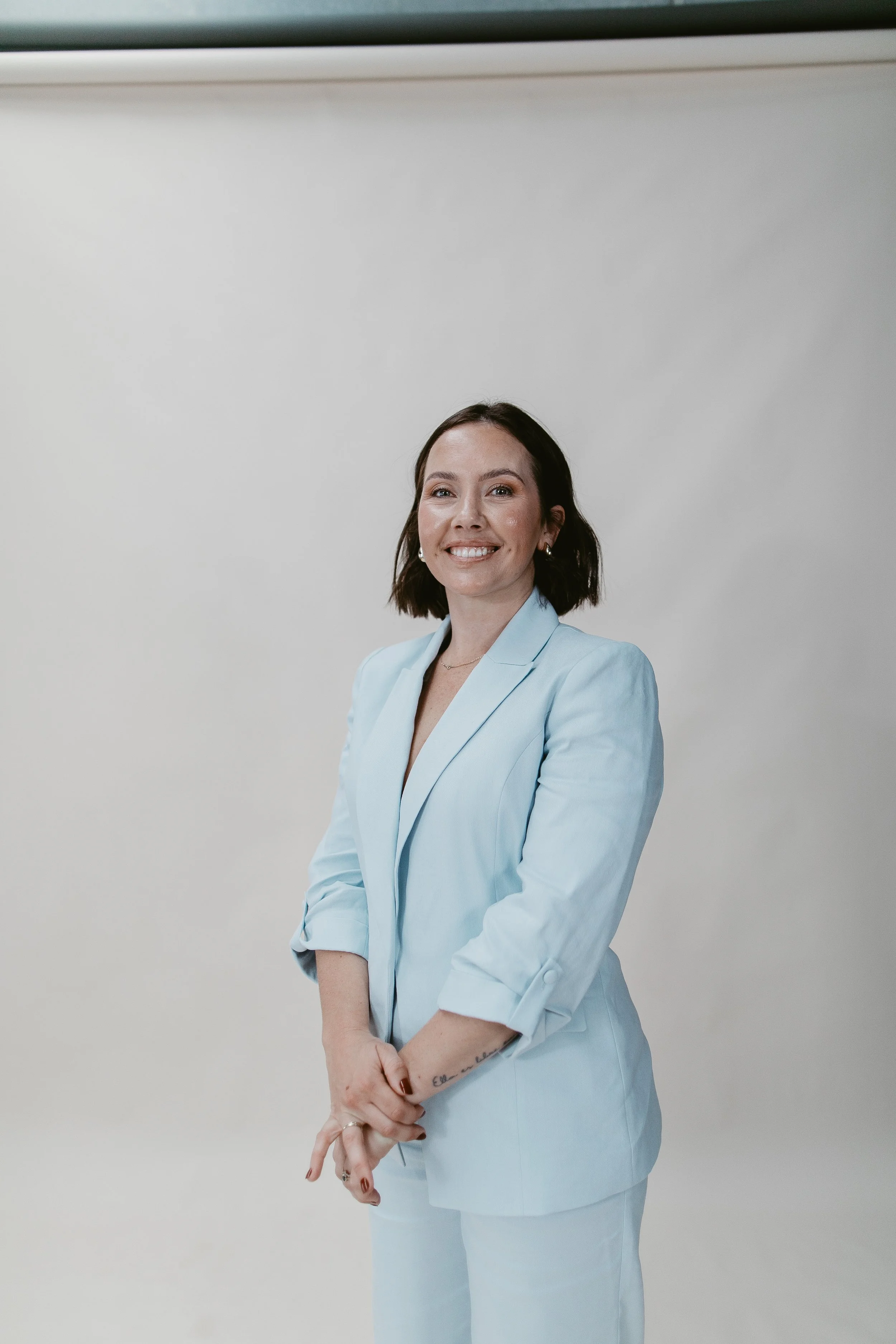 A woman with shoulder-length dark hair dressed in a light blue blazer and matching pants, standing against a plain white background. She is smiling, showing her teeth, with her arms crossed in front of her, her right hand gently holding her left wrist, and her left arm resting across her stomach.
