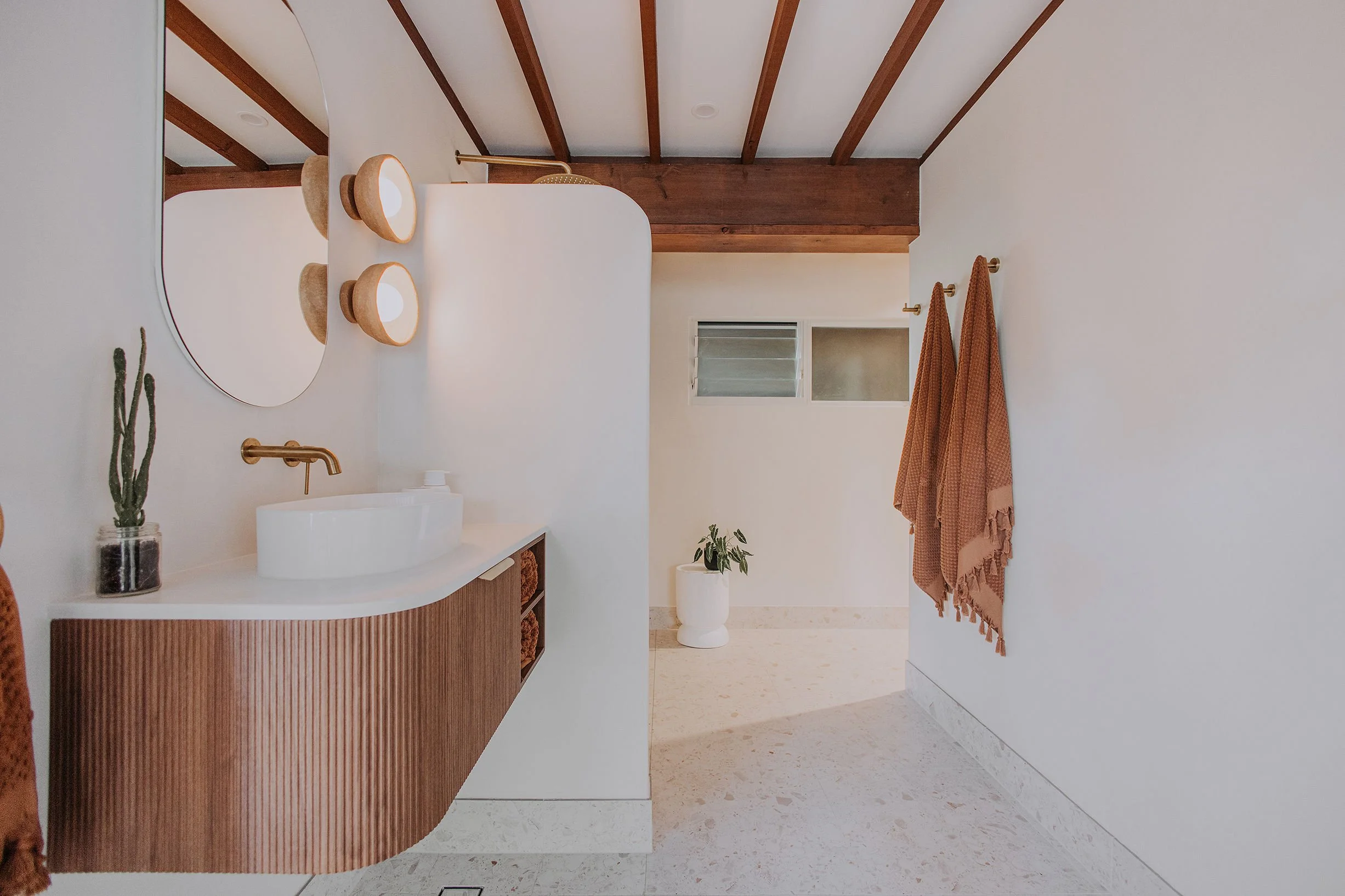 A modern bathroom with a curved wooden vanity, white vessel sink, and gold faucet. Two beige wall sconces above the mirror, a small cactus in a glass jar, a potted plant on a white stand, and beige towels hanging on gold hooks. The room has white walls, a window with horizontal blinds, and exposed wooden ceiling beams.