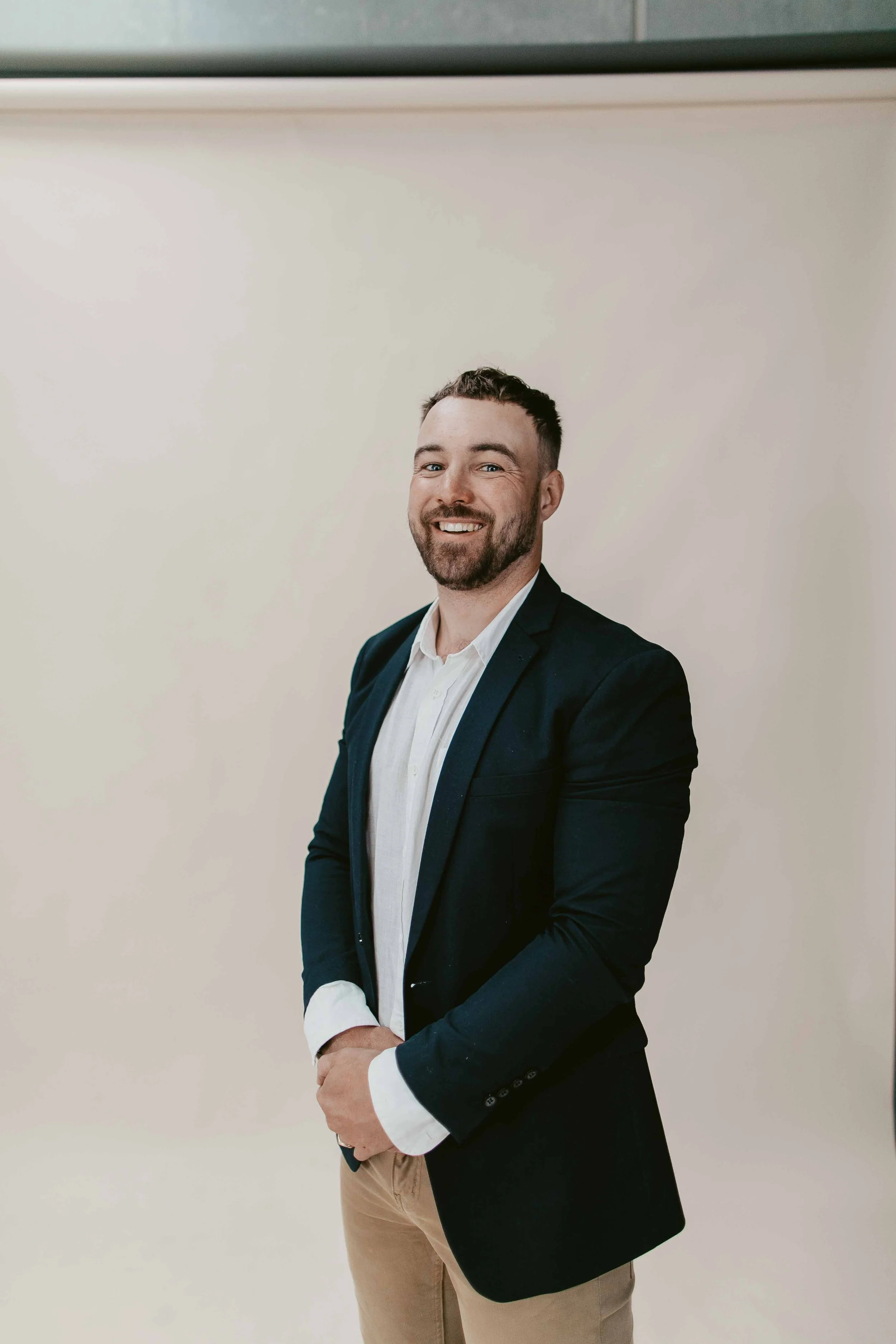 A man with a beard and short dark hair, dressed in a white shirt and black blazer, smiling and standing against a plain white wall.