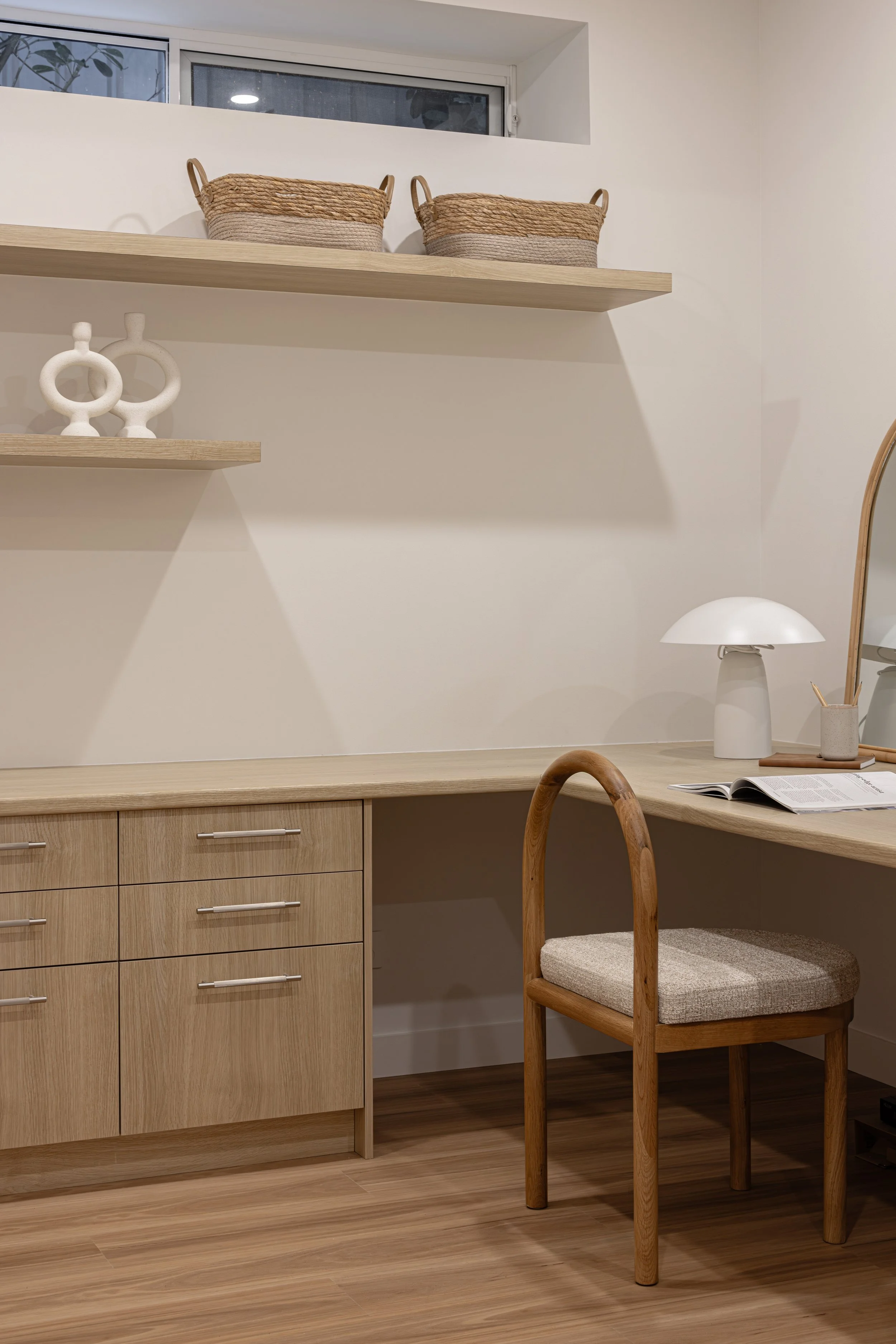 A home office with wooden drawers, a wooden chair with a cushioned seat, a desk lamp, an open book, and wall-mounted wooden shelves holding decorative baskets and sculptures.