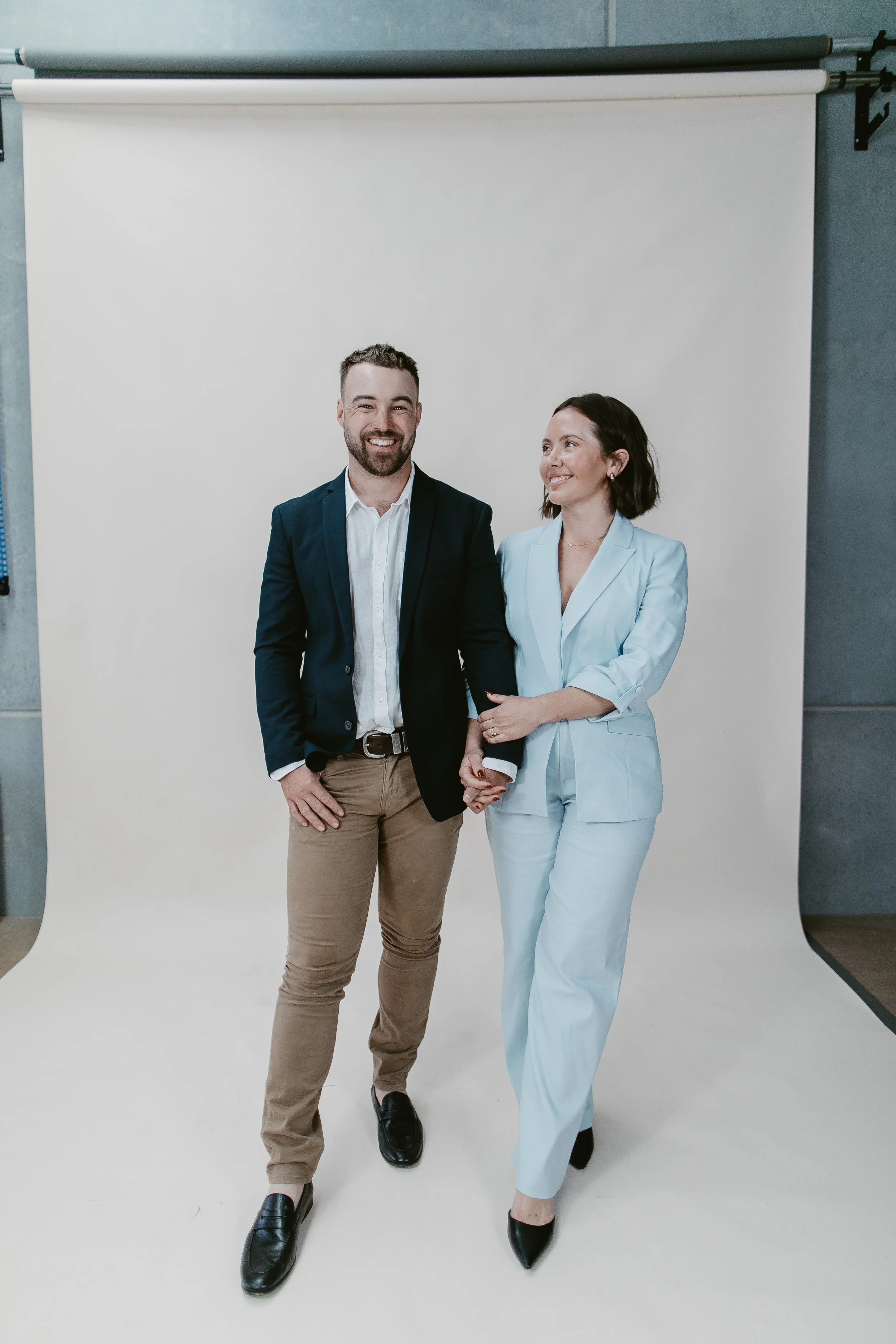 A man and a woman standing together in front of a plain white backdrop, holding hands, dressed in formal attire. The man is wearing a navy blazer, white shirt, khaki pants, and black shoes. The woman is wearing a light blue pantsuit and black heels.