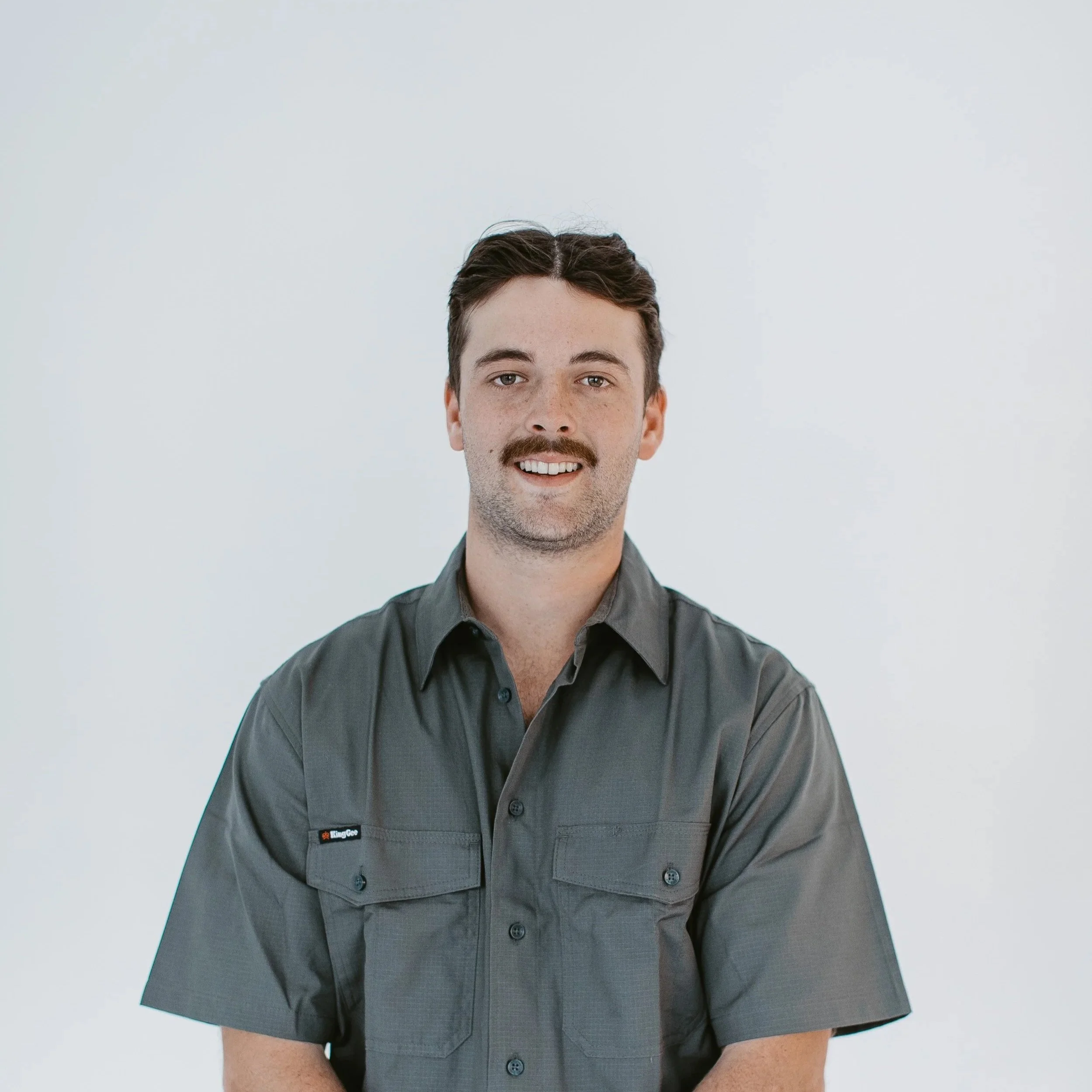 Close-up photo of a man with dark hair, mustache, and beard, wearing a dark gray shirt, smiling, standing against a plain light gray background.