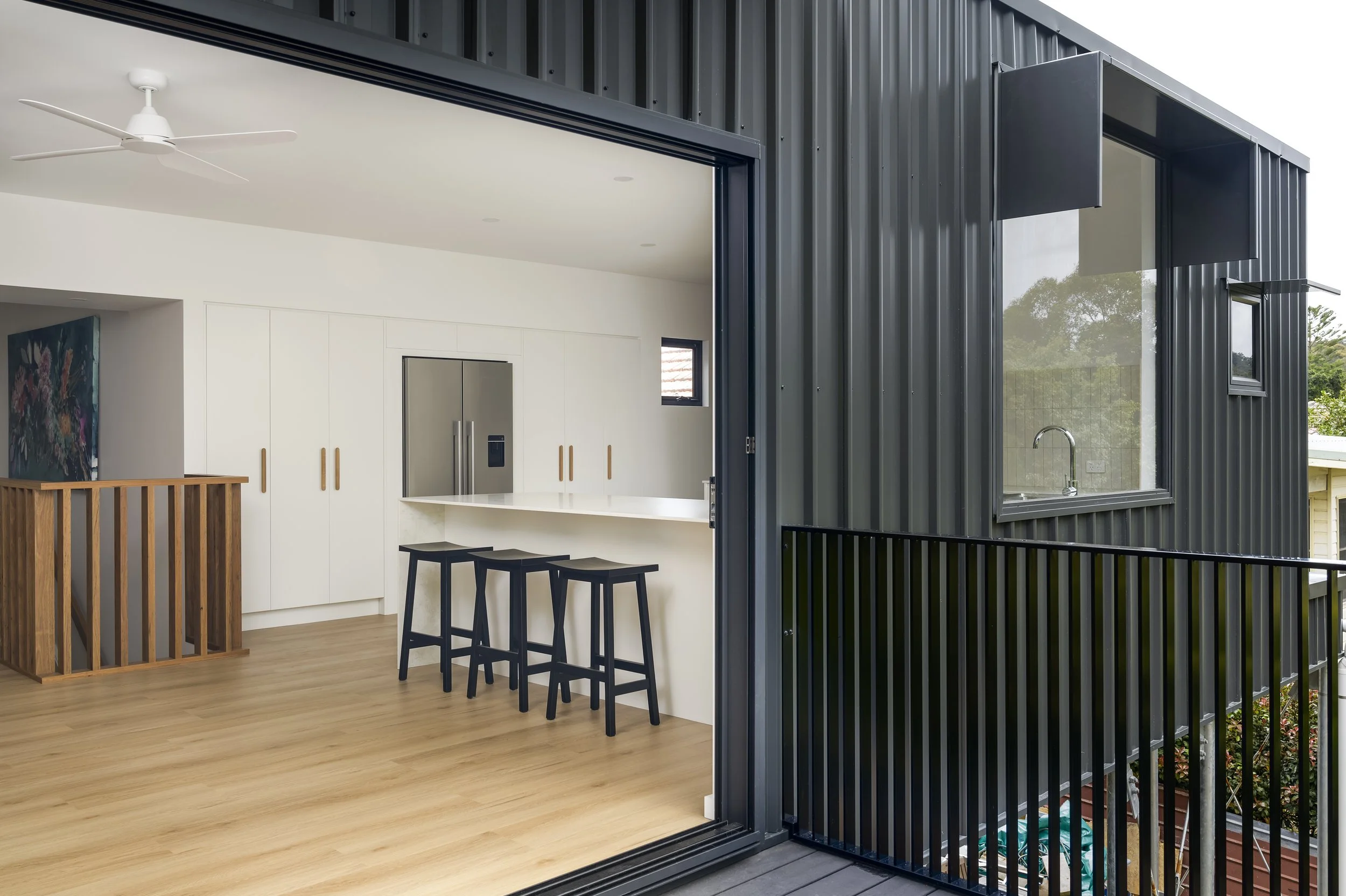 Interior view of a modern home with a balcony, black metal exterior wall, and a sliding glass door revealing a bright living space with white walls, a ceiling fan, wooden flooring, and a kitchen island with stools.