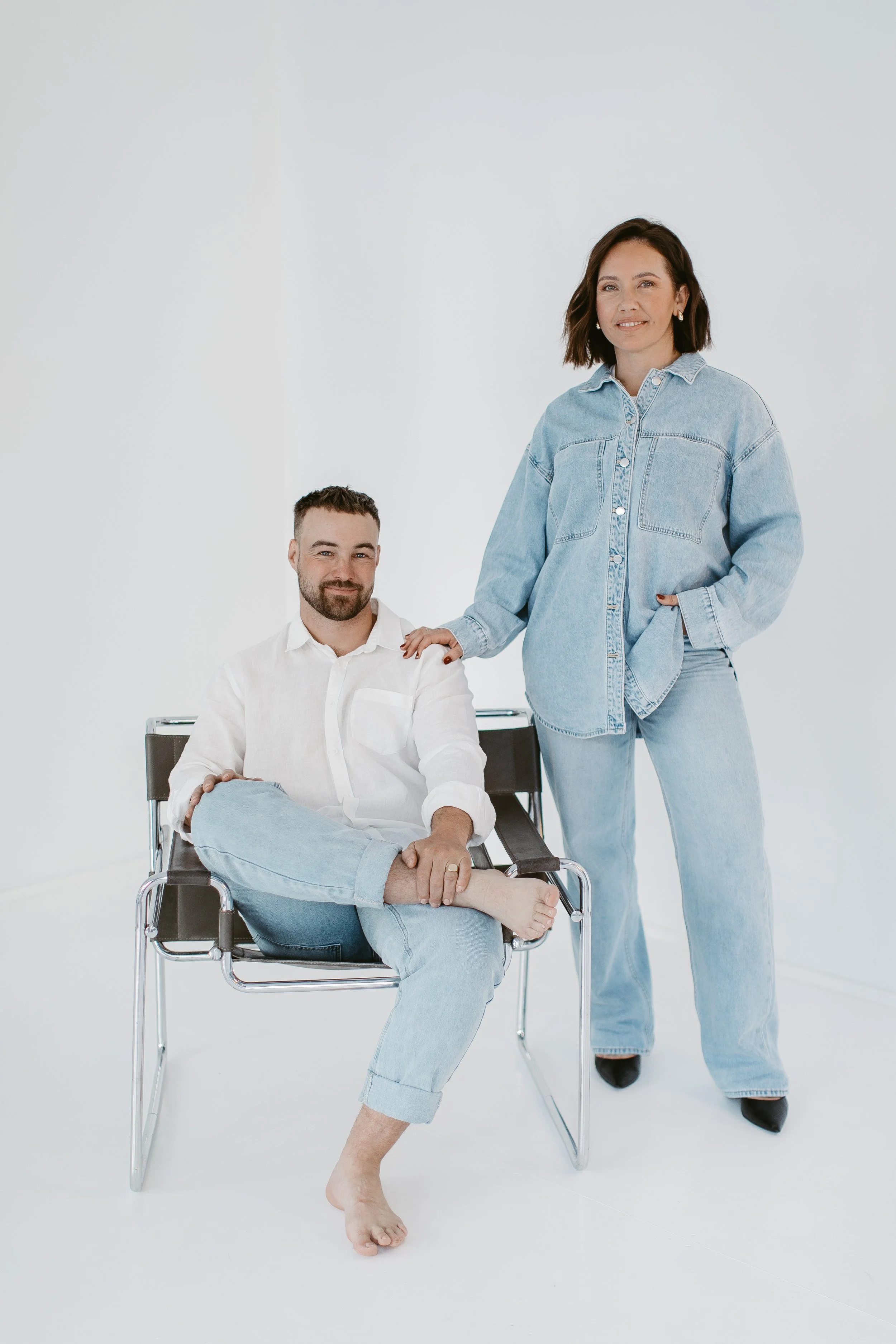 A woman in casual denim clothing standing and a man in casual clothing sitting in a chair in a plain white room.