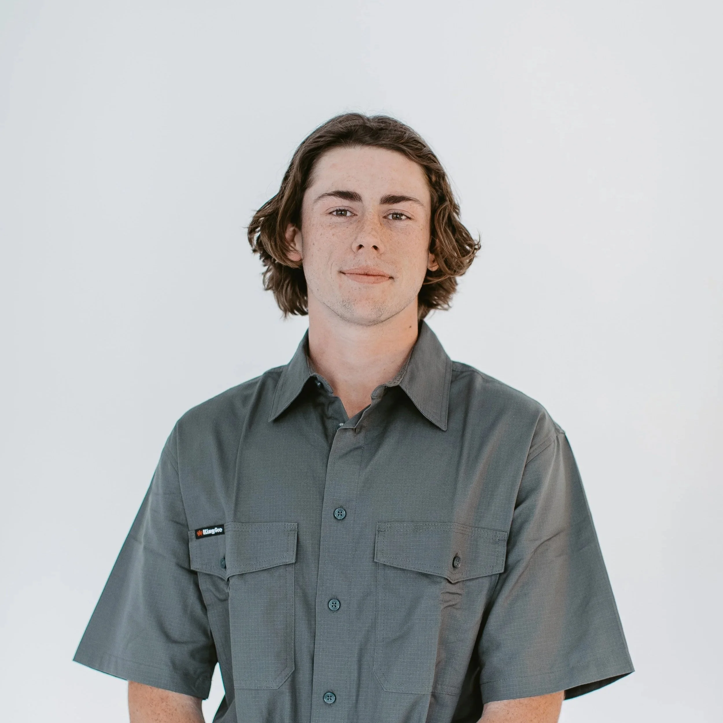 A young man with medium-length brown hair, wearing a gray uniform shirt with a pocket on the left side labeled 'KingGee', standing against a plain, light-colored background.