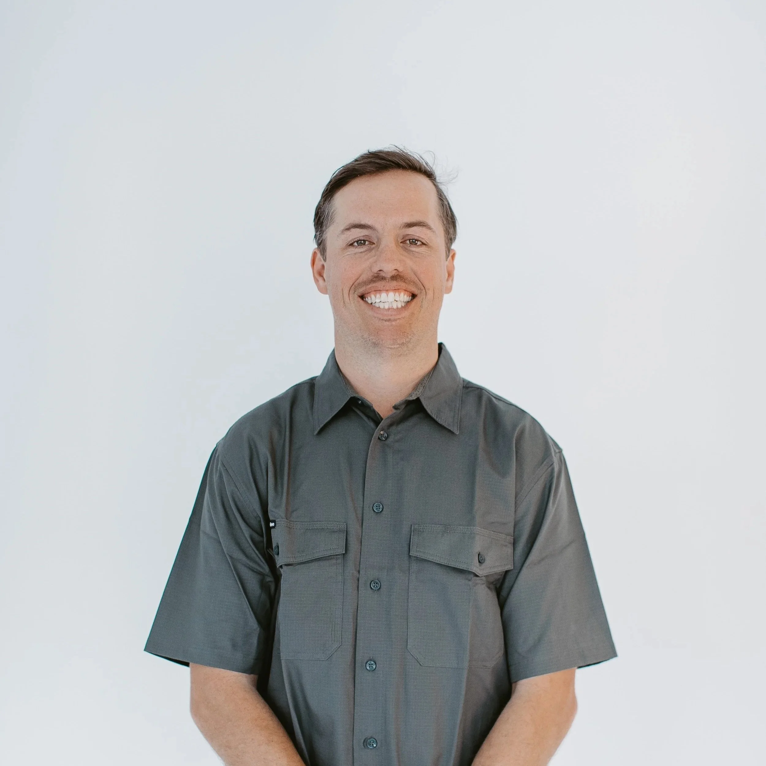 A smiling man with short brown hair, wearing a dark gray button-up shirt, standing against a plain white background.