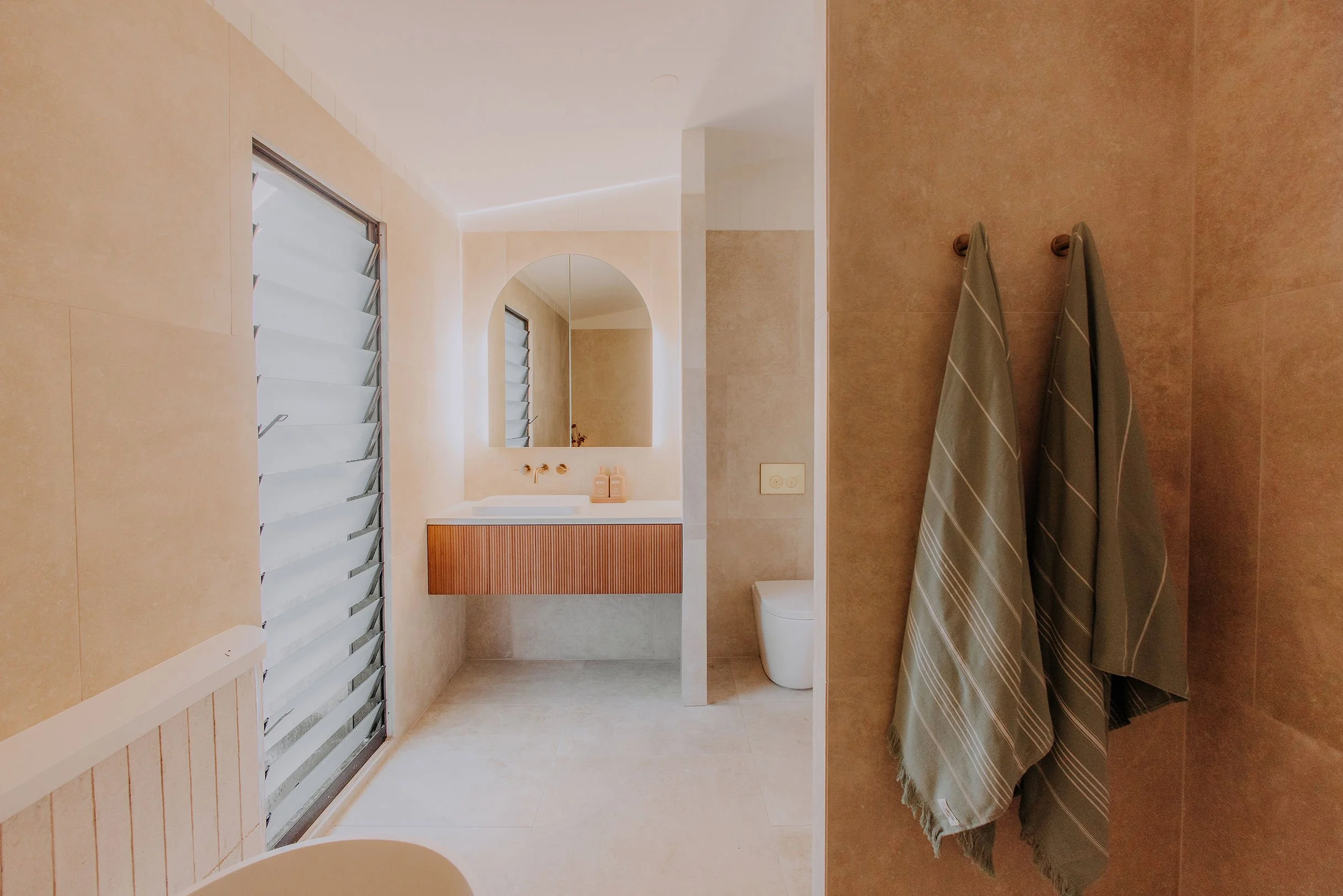 A modern bathroom with beige tiled walls and floors, a white sink with a mirror above, a toilet, and a window with adjustable blinds. There are two towels hanging on hooks to the right.