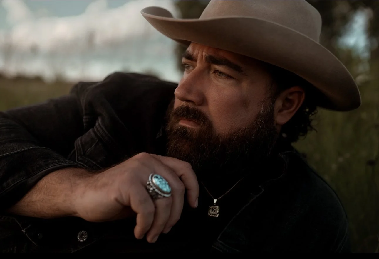 Man with a beard wearing a cowboy hat, black shirt, and ring, resting his chin on his hand, outdoors with trees in the background.
