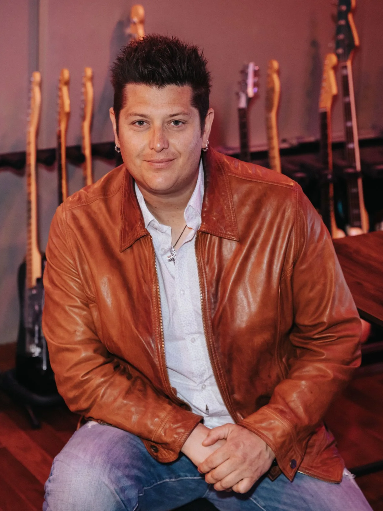 A person with short dark hair, earrings, and a slight smile, sitting indoors with multiple guitars hanging on the wall behind them.
