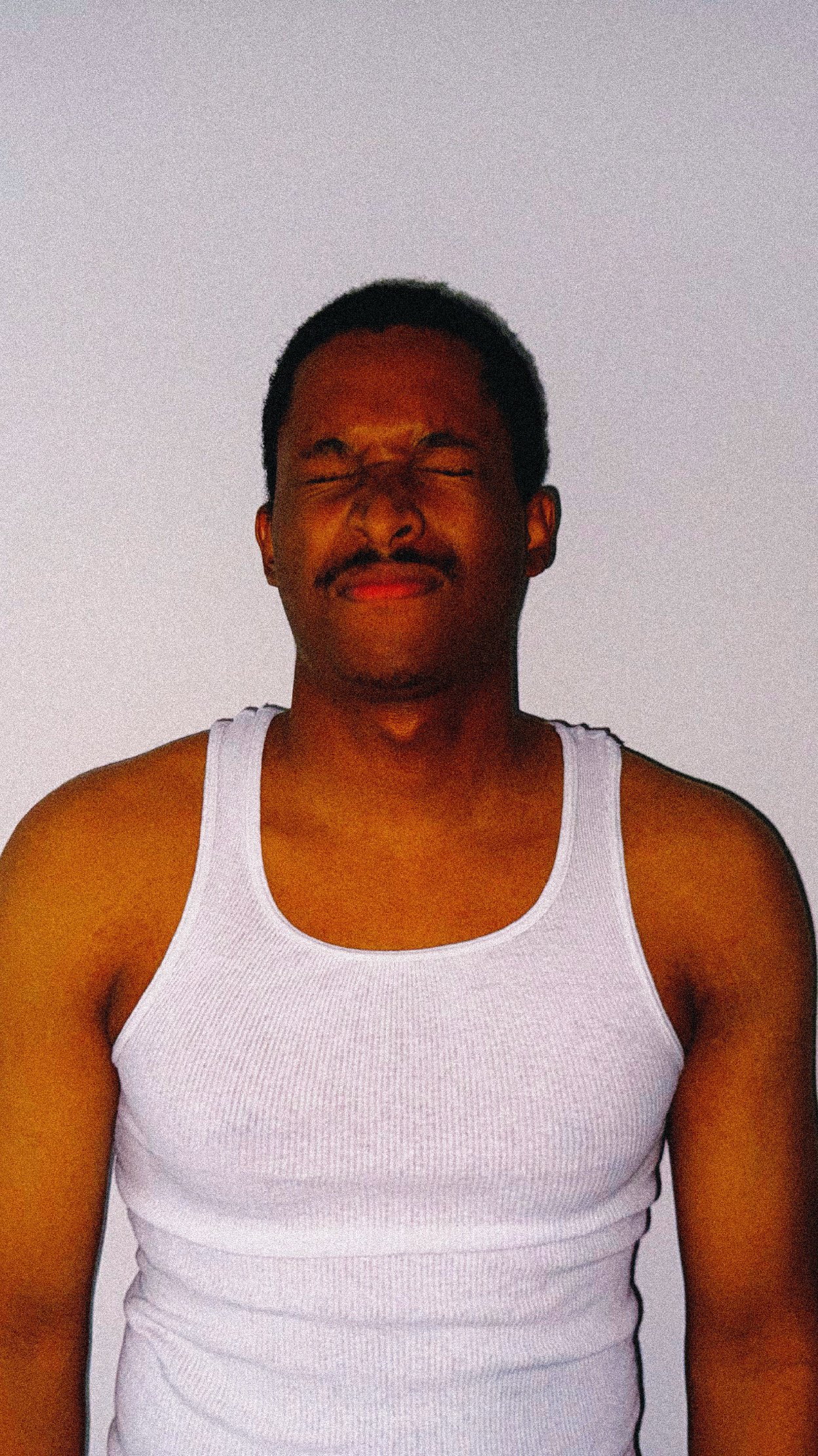 A person with medium-dark skin tone, short black hair, wearing a white tank top, with eyes closed and a slight smile, standing against a plain, light-colored background.