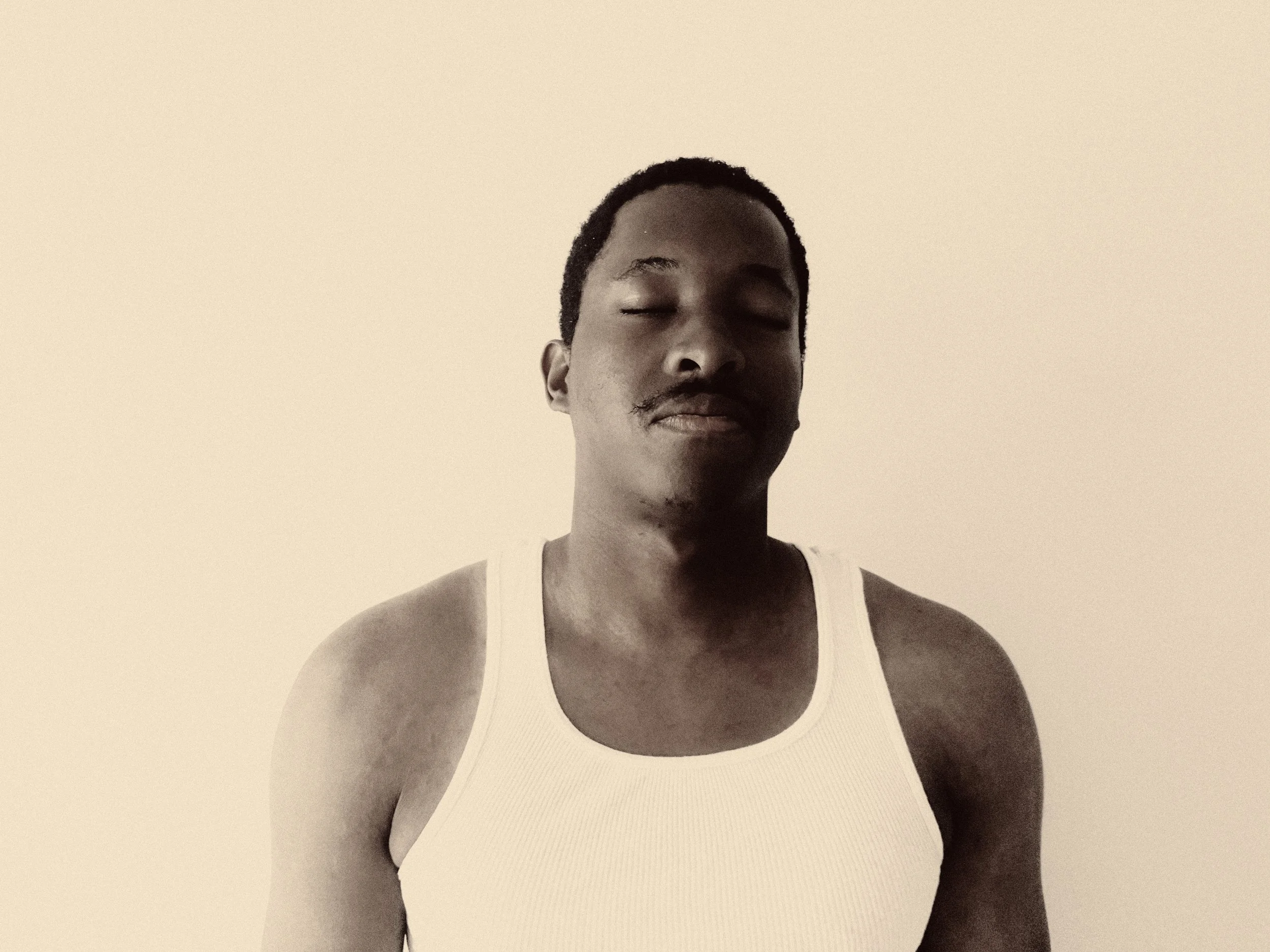 A black-and-white photo of a man with closed eyes, wearing a white tank top, and short curly hair, against a plain background.
