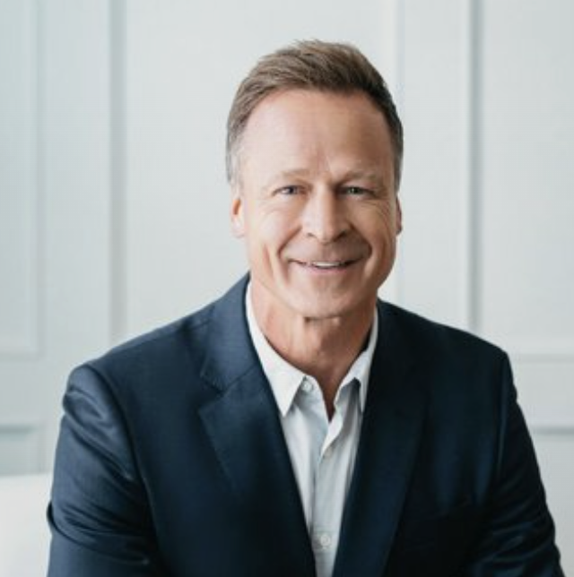 Headshot of a smiling middle-aged man in a dark suit and white shirt, sitting in front of a neutral background.