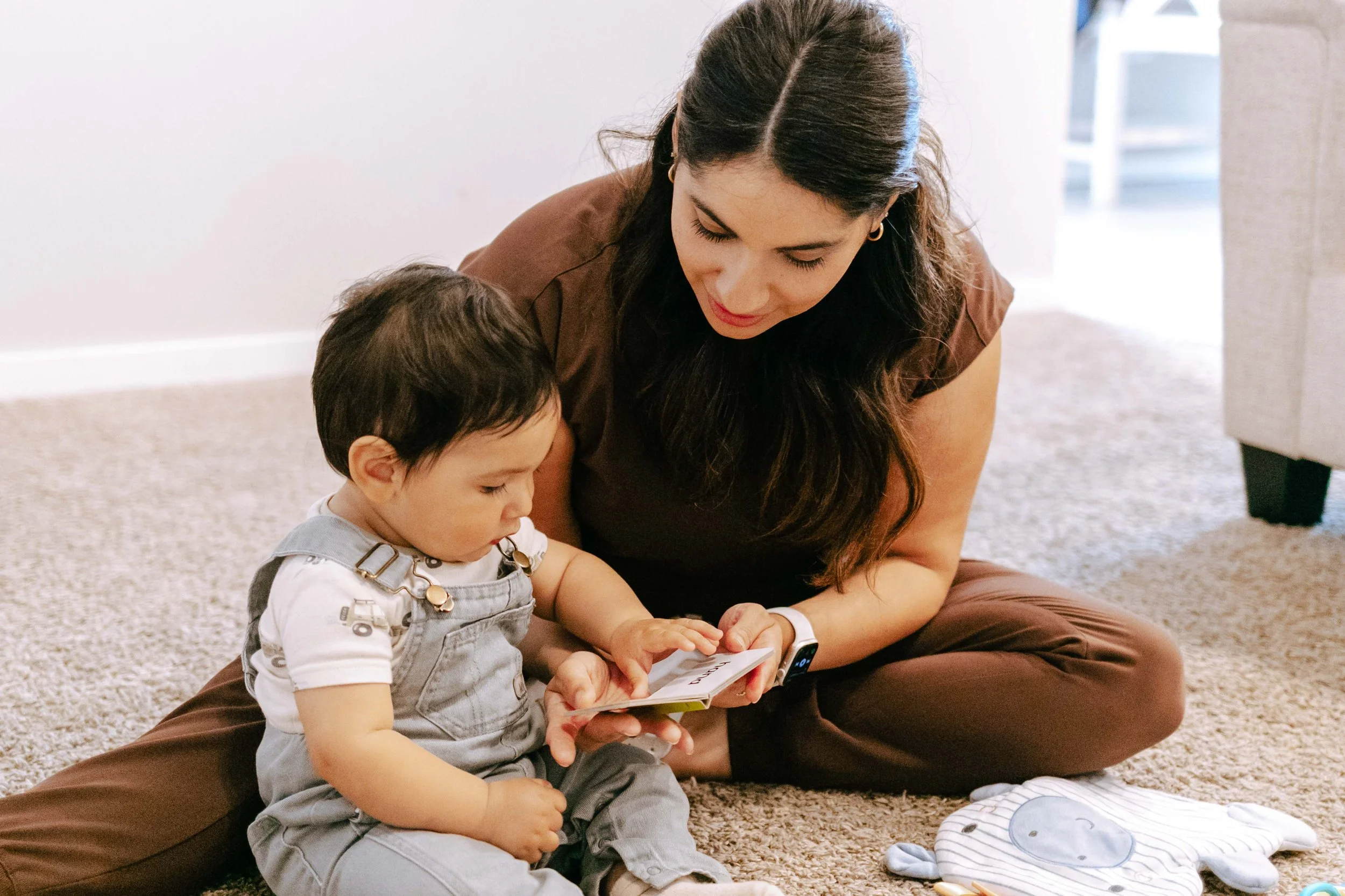 A woman and a young boy sitting on a beige carpeted floor, looking at a book or card together. The woman has long dark hair and is wearing a brown. The boy has dark hair, is wearing a white shirt with gray overalls, and is focused on the item they are holding.
