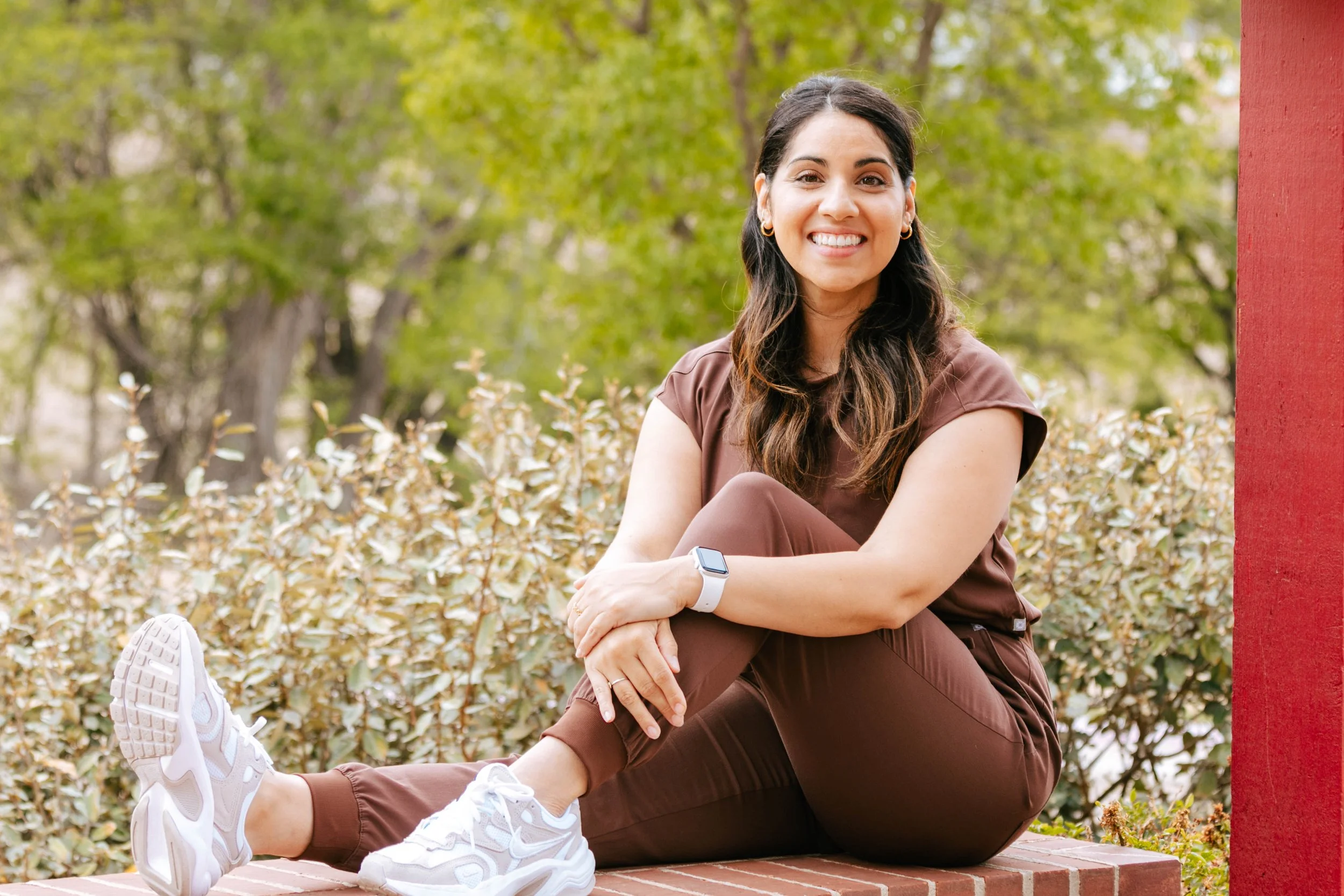 A woman with long dark hair, smiling and sitting outdoors on a brick surface next to a red pillar, with greenery and trees in the background.