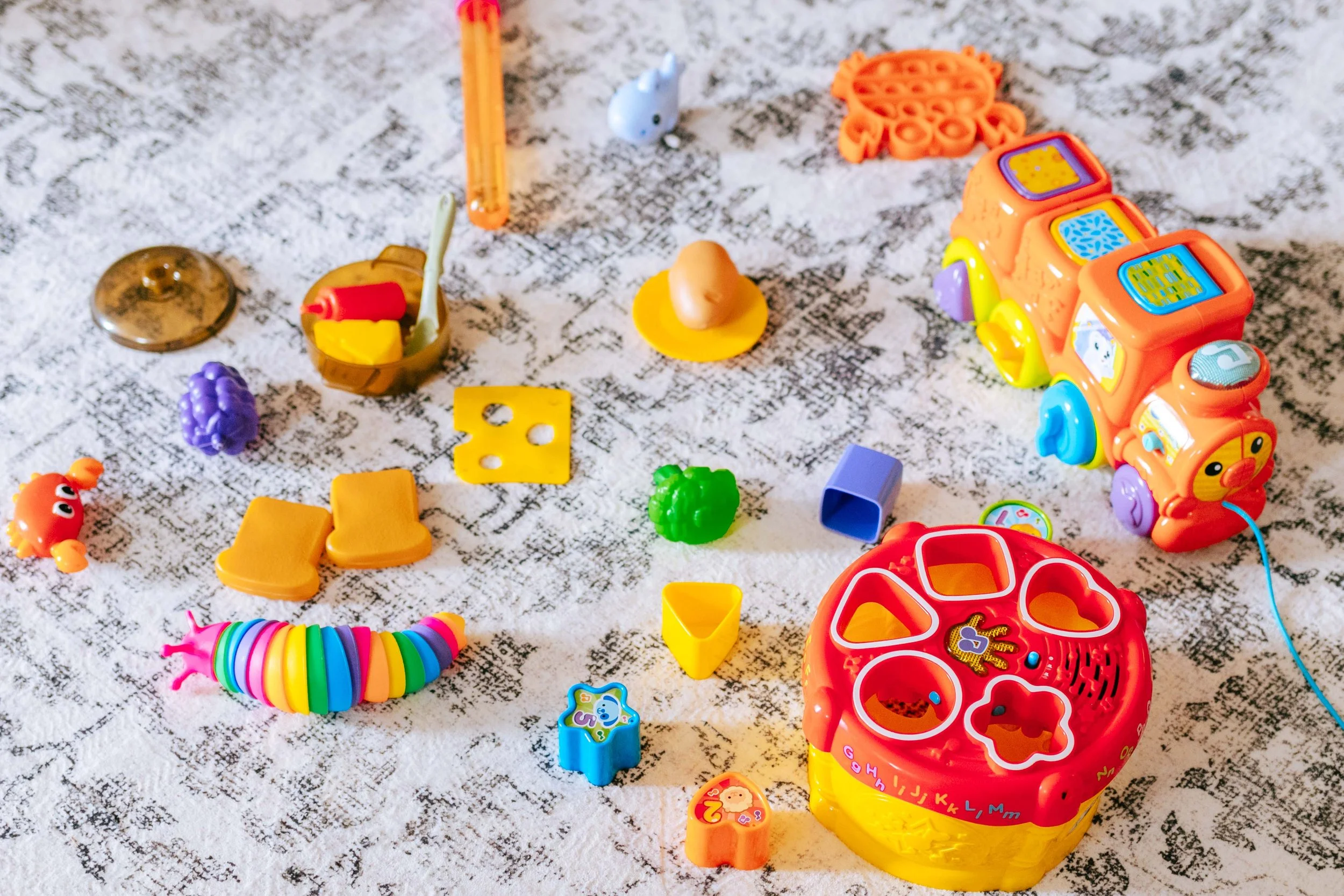Colorful children's toys spread out on a white textured surface, including a rainbow-colored unicorn, a yellow and orange shape sorter, a plastic train with animal faces, a small white figurine, and various small blocks and accessories.