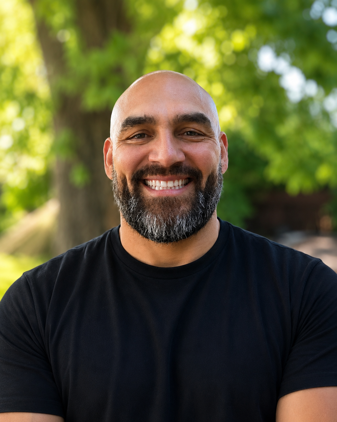 A smiling man with a beard and shaved head outdoors with a background of green trees.