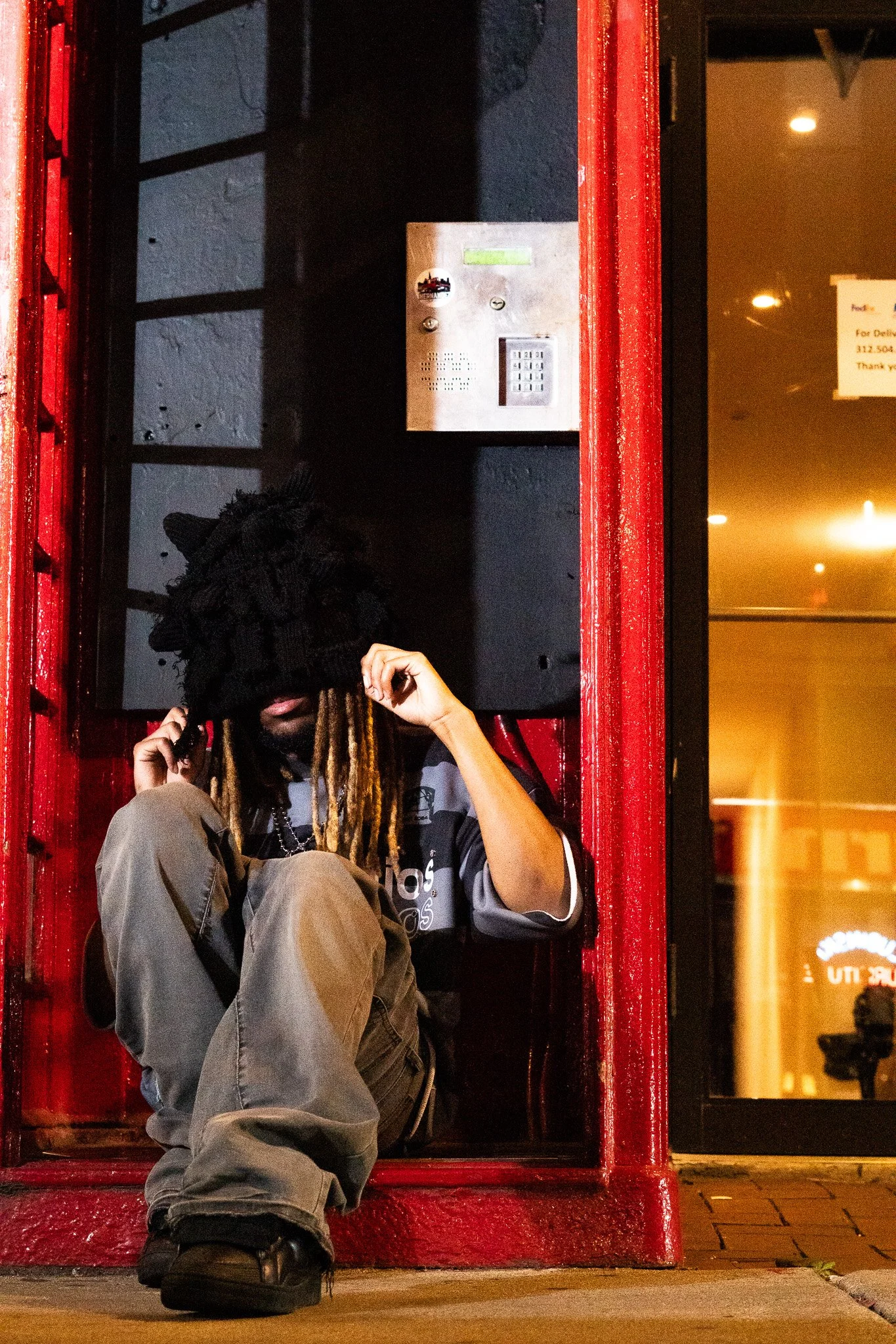 A person with dreadlocks, black face mask, black and gray Adidas jacket, beige cargo pants, and black boots sitting on the sidewalk outside a red phone booth at night, with a phone in hand.