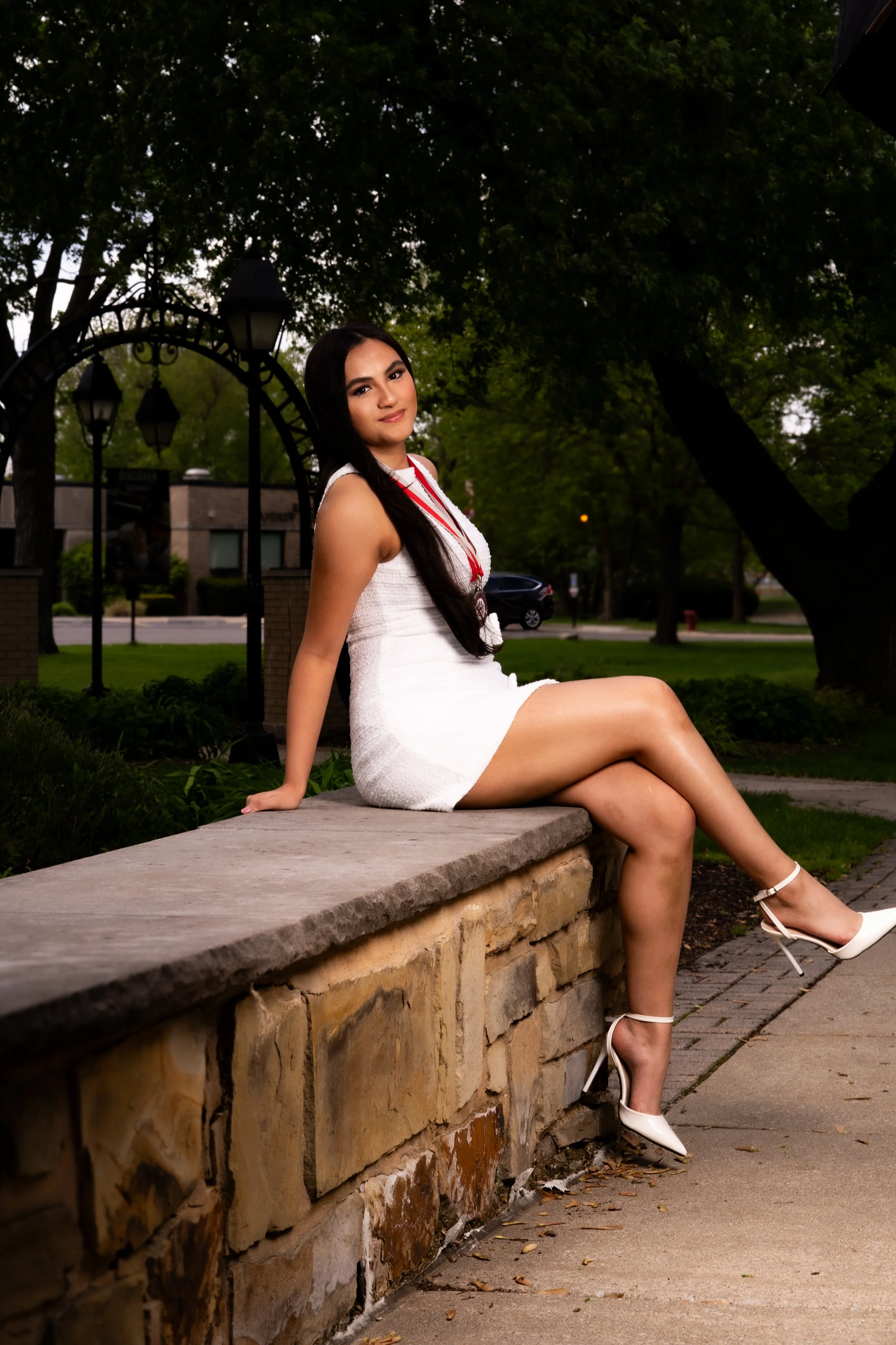 A woman in a white dress and high heels sitting on a stone ledge in a park with green trees and a street with a car in the background.