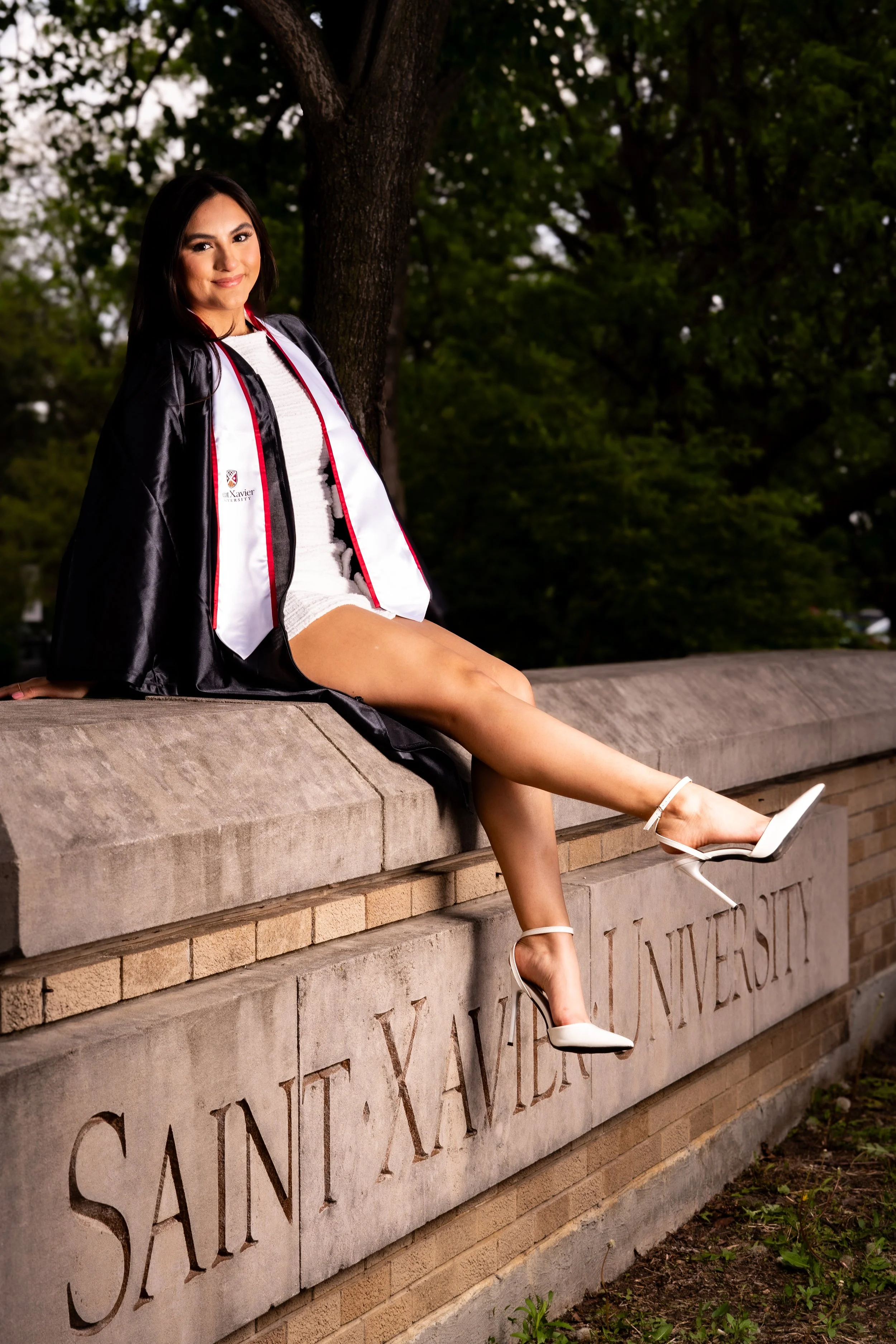 A young woman in a graduation gown and white dress, sitting on a stone wall outside with trees in the background. She is wearing white high heels and smiling.