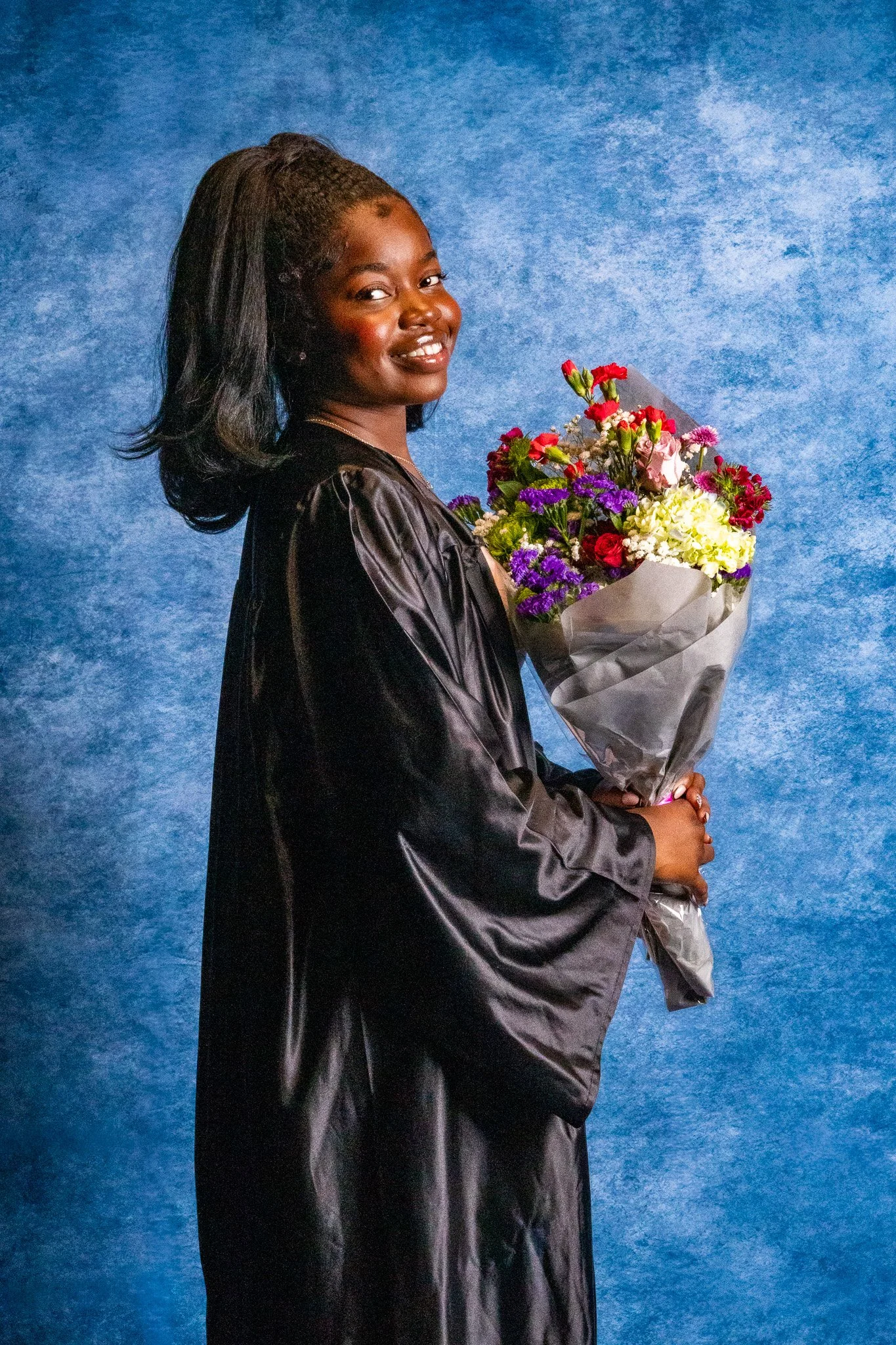 A young woman with dark skin, black wavy hair, and a bright smile, wearing a black graduation gown, holding a colorful bouquet of flowers, standing against a blue textured background.