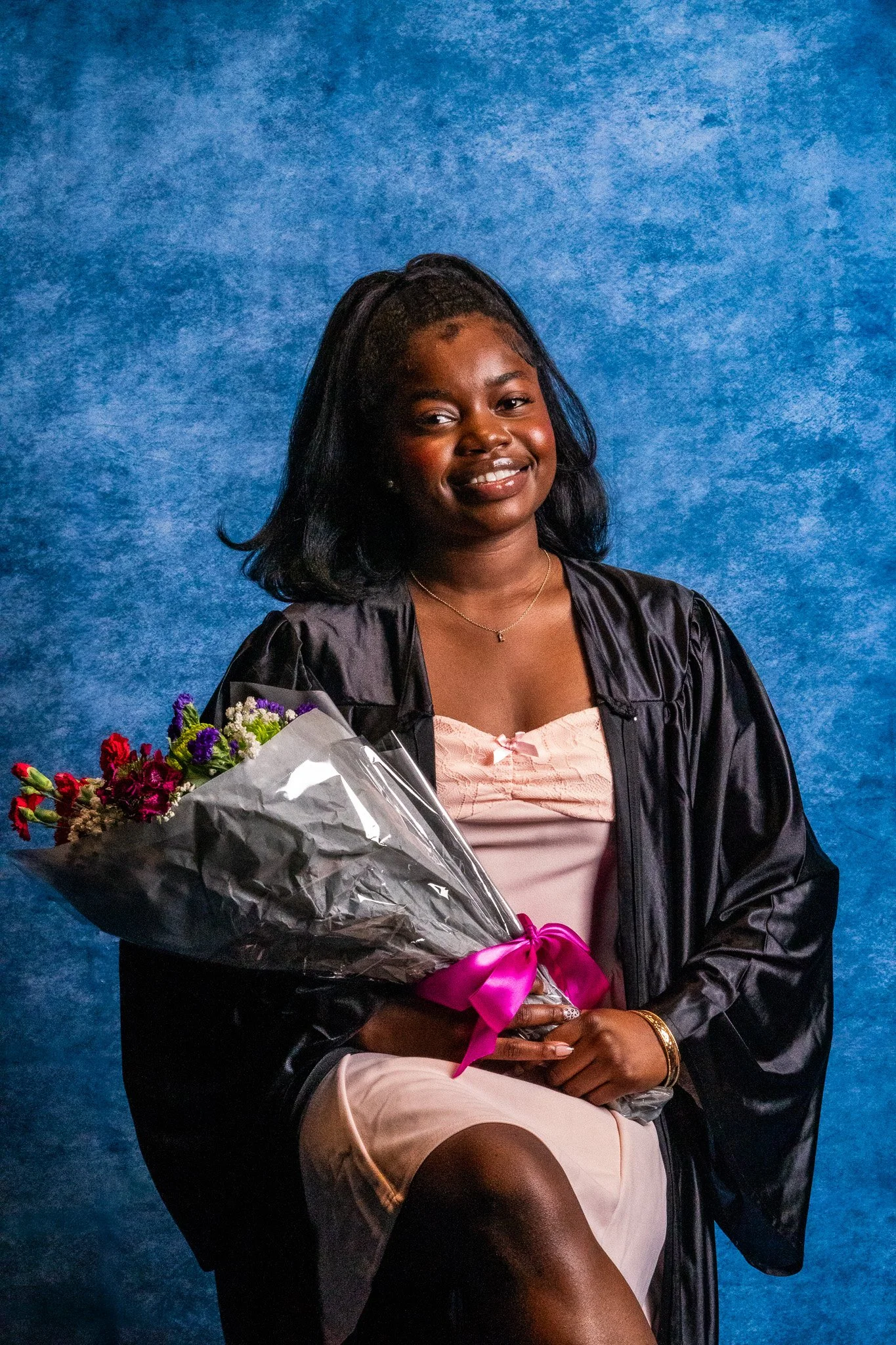 A young woman in a graduation gown and cap holding a bouquet of flowers, sitting against a blue background.